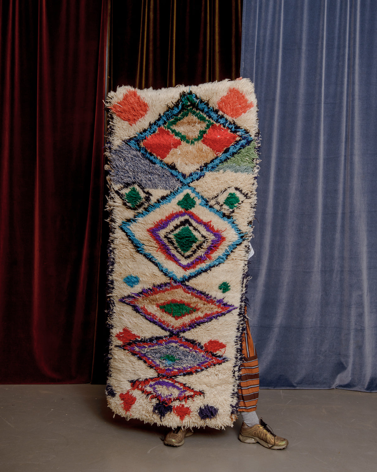 Vintage Berberlin boucherouite runner rug being held up in front of a velvet curtains.