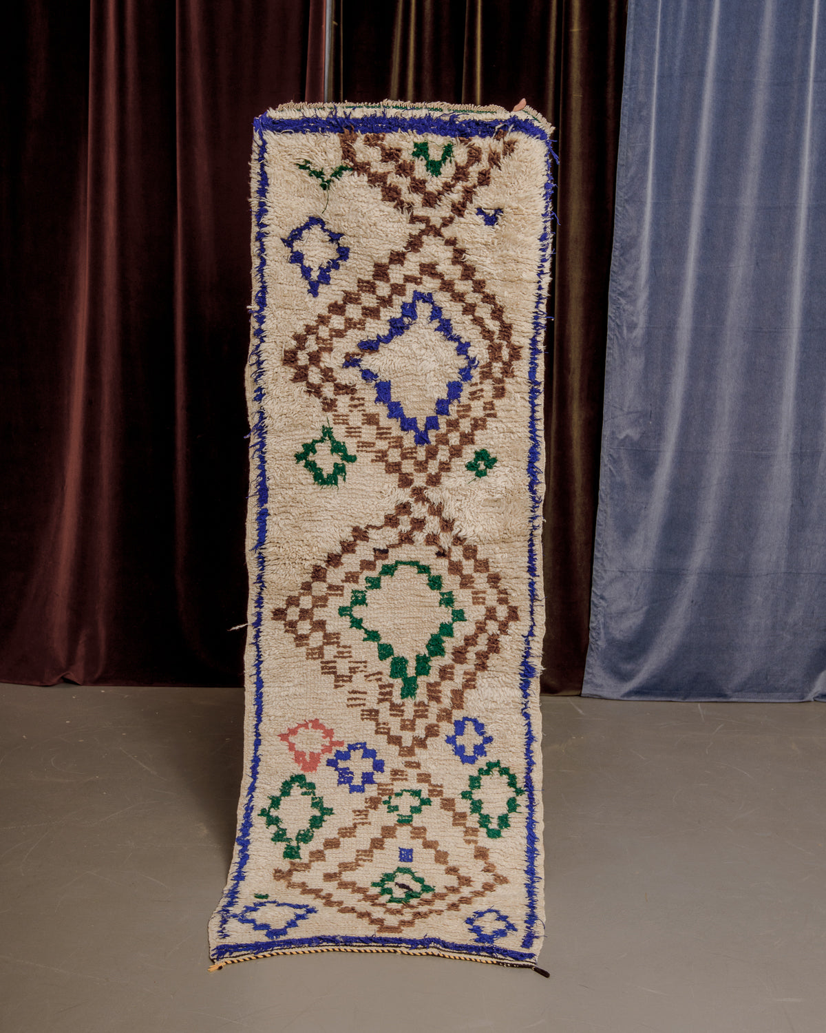 Vintage Berberlin boucherouite runner rug being held up in front of a velvet curtains.