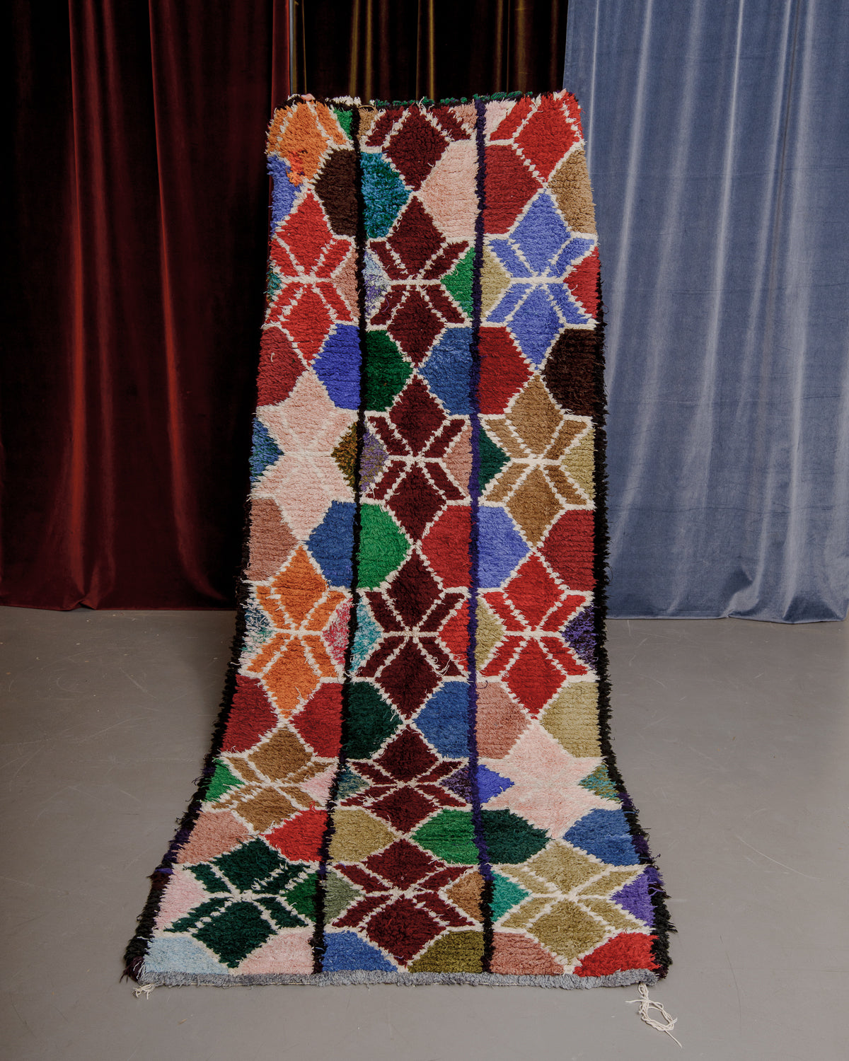 Vintage Berberlin boucherouite runner rug being held up in front of a velvet curtain. 