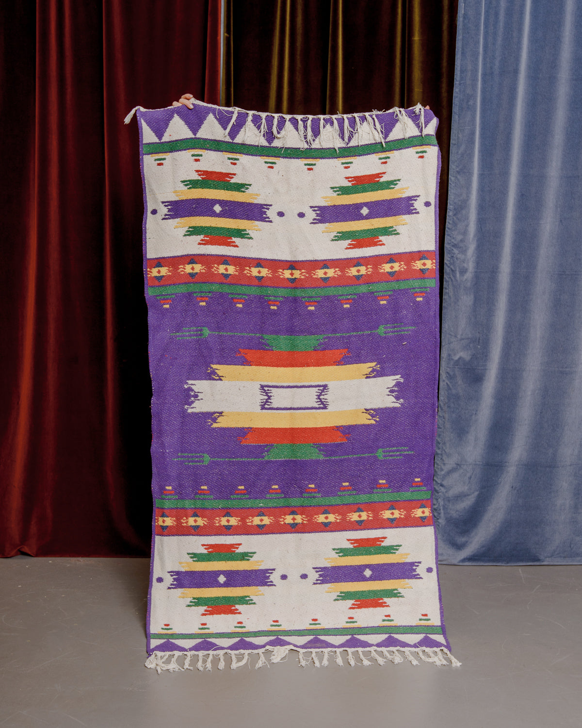 Vintage Berberlin boucherouite kelim runner rug being held up in front of a velvet  curtains.