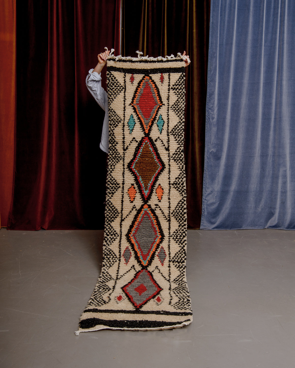 Vintage Berberlin boujaad runner rug being held up in front of a velvet curtains.