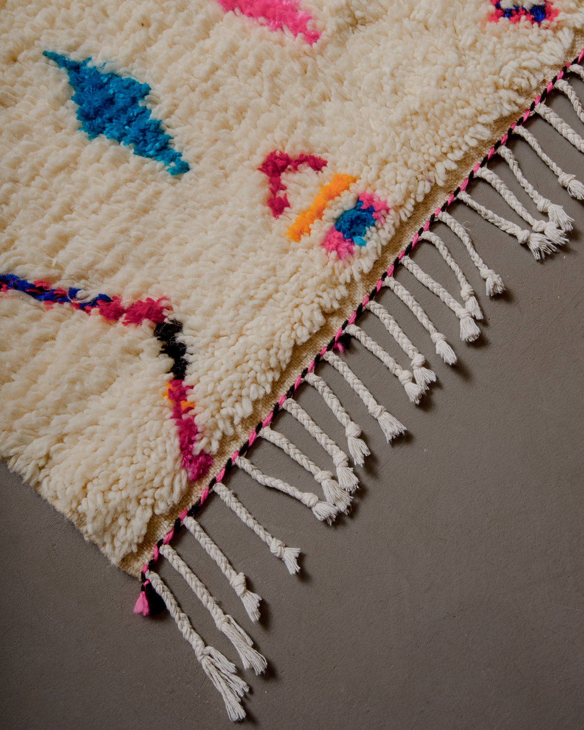 Corner of azilal rug with white cotton tassels and pink and black edge.