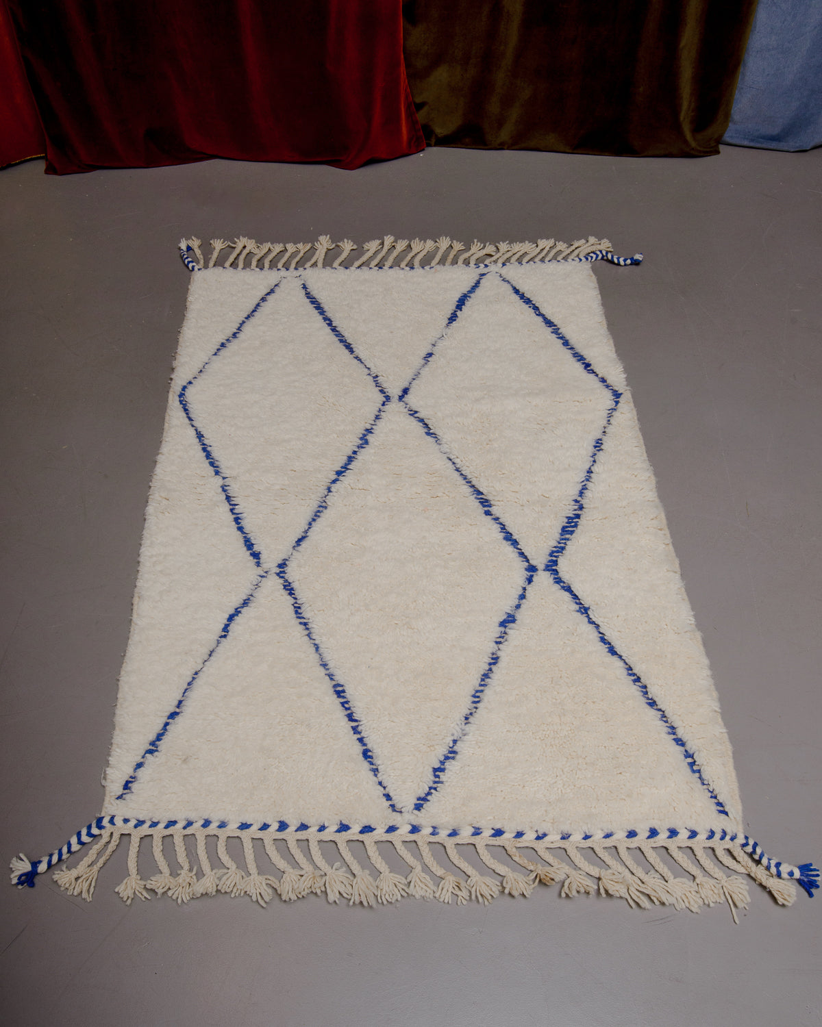 Berberlin Beniourain rug with blue diamond pattern on grey floor in front of velvet curtains. 