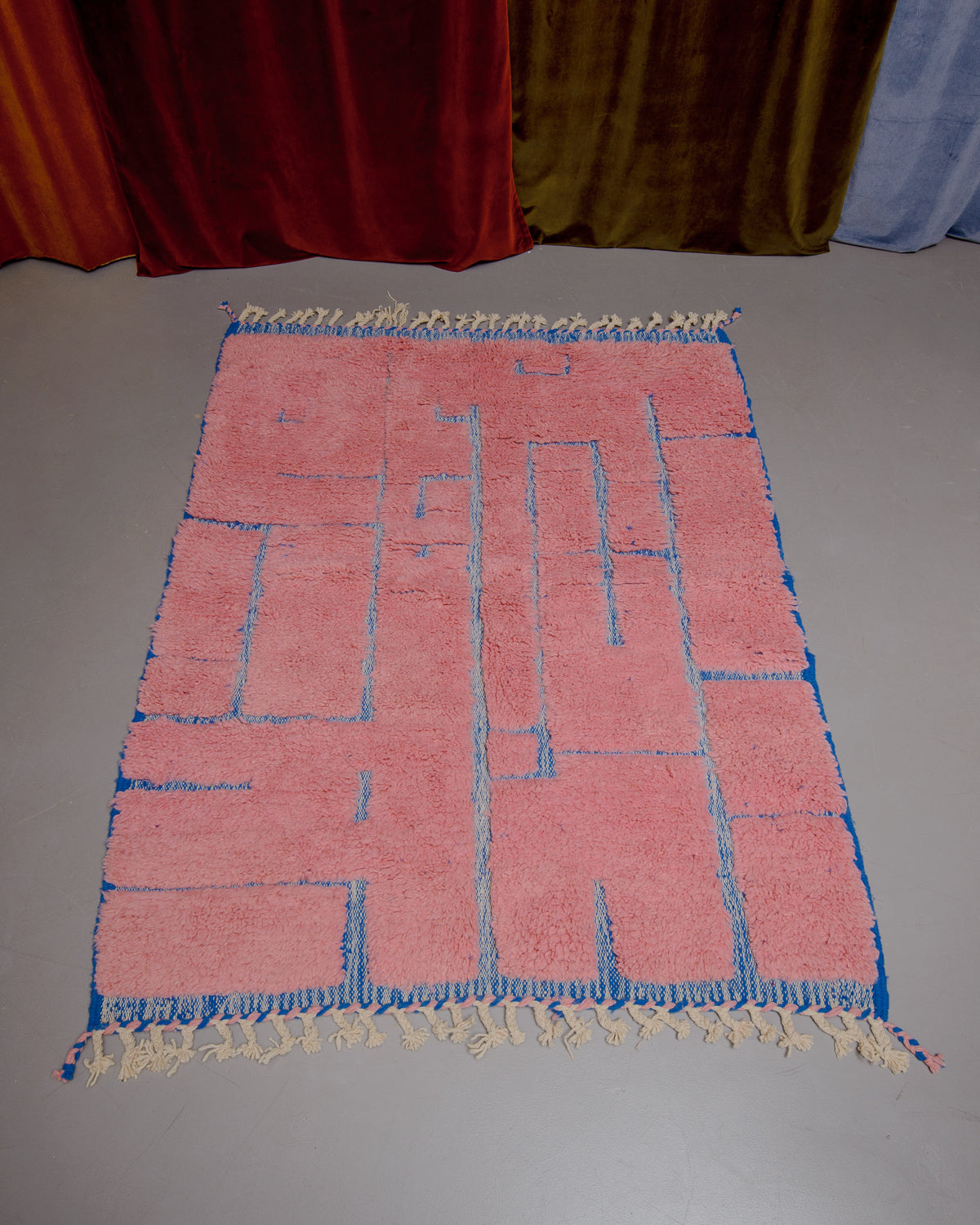 Beniourain rug in pink and blue wool on grey floor in front of velvet curtains. 