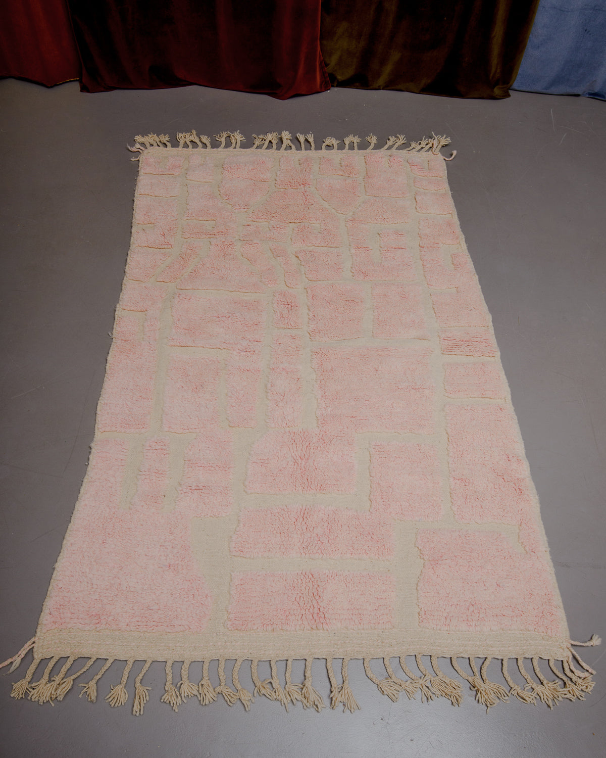 Beniourain rug in pale rose and natural wool on grey floor in front of velvet curtains. 