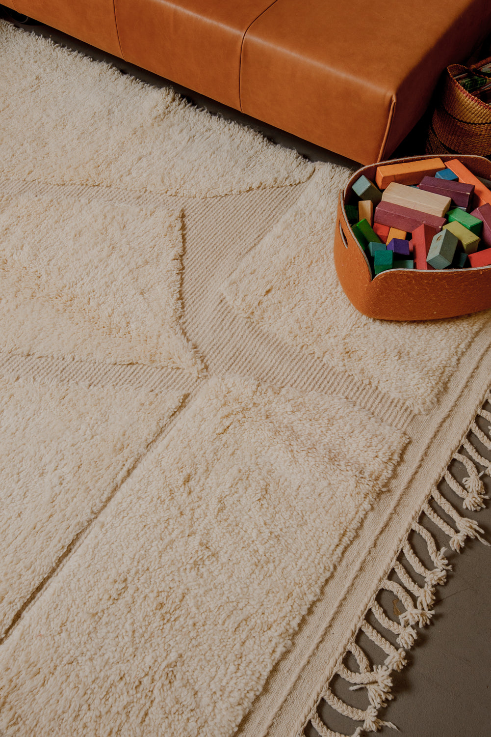 Interior photo of cream wool rug in a setting with leather couch in cognac and a basket of colourful wooden blocks.