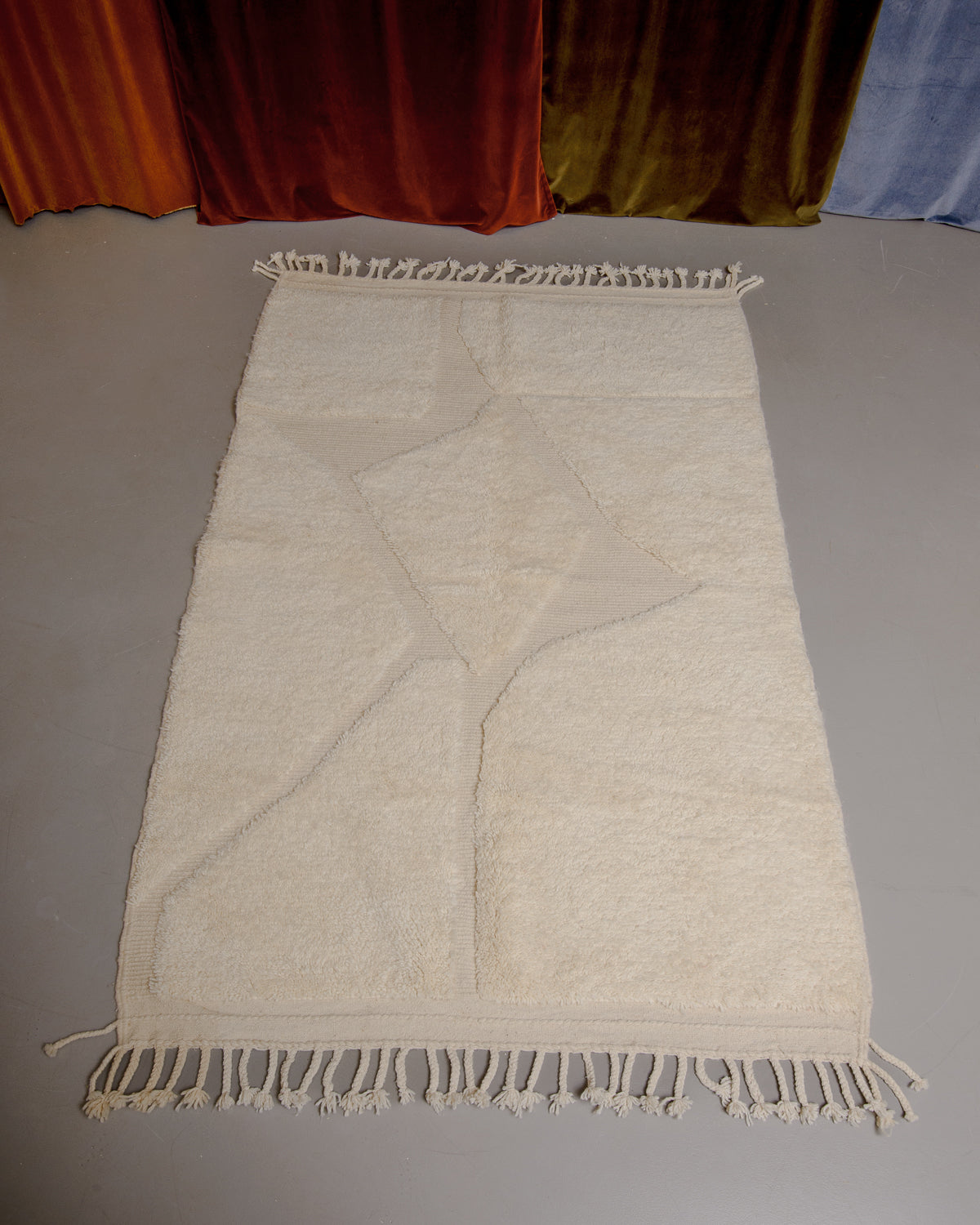 Cream beniourain rug with embroidered design on grey floor in front of velvet curtains. 