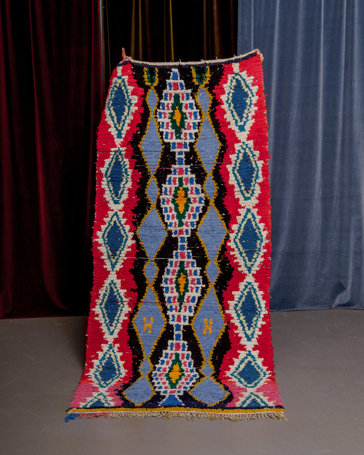 Vintage Berberlin boucherouite runner rug being held up in front of a velvet curtain. 