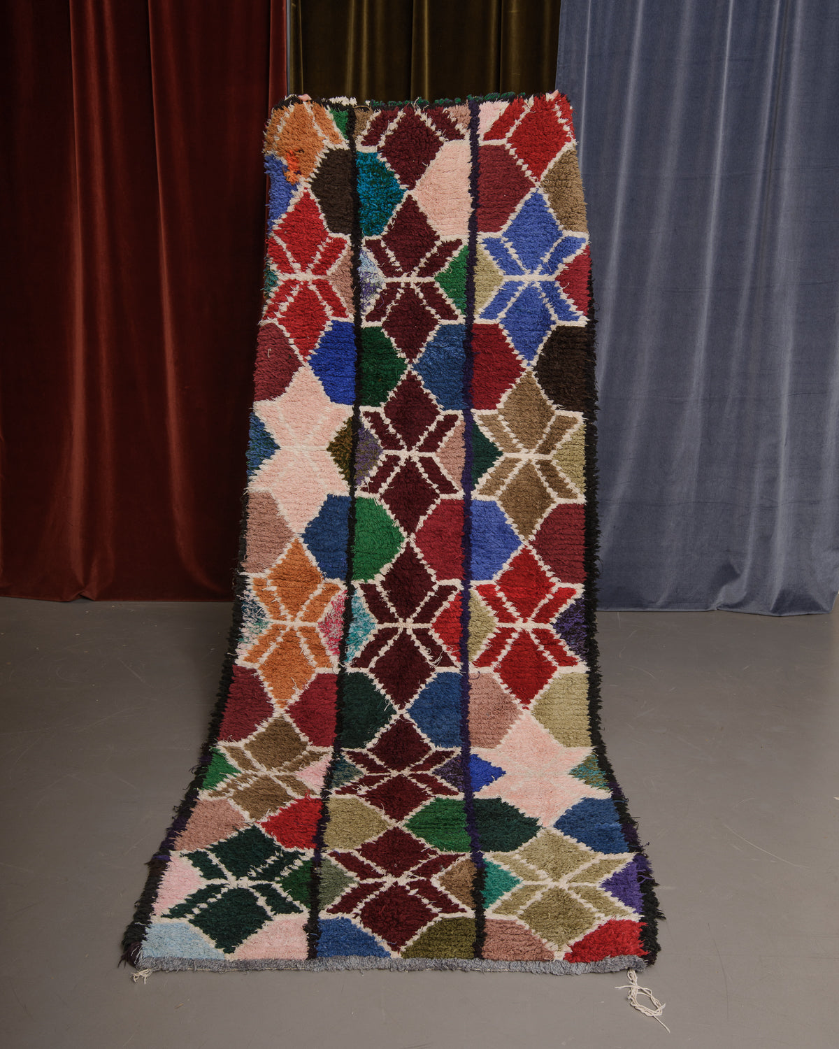 Vintage Berberlin boucherouite runner rug being held up in front of a velvet curtain. 