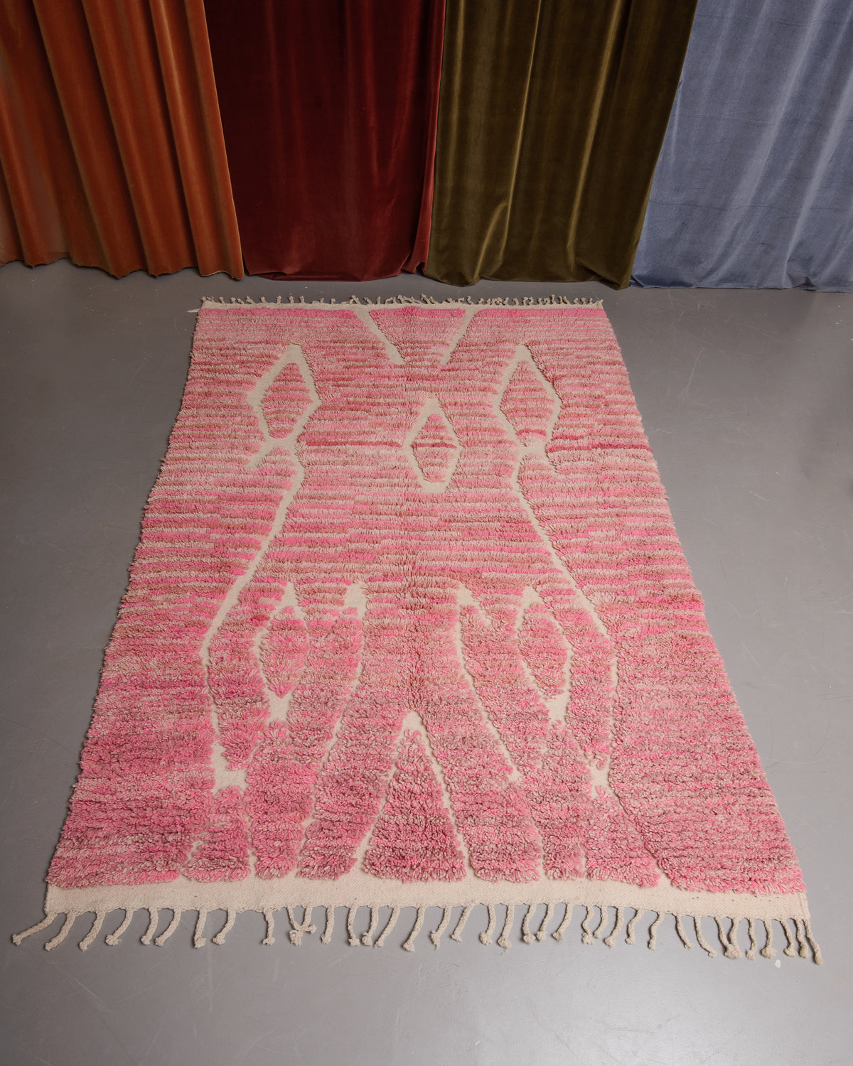 Berberlin Boujaad rug in different shades of pink wool with organic woven lines on grey floor in front of velvet curtains.