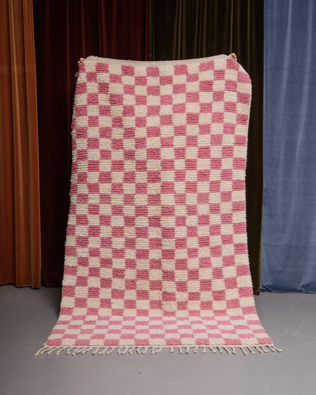 Berberlin Boujaad rug in pink and cream checkered wool being held in front of velvet curtains.