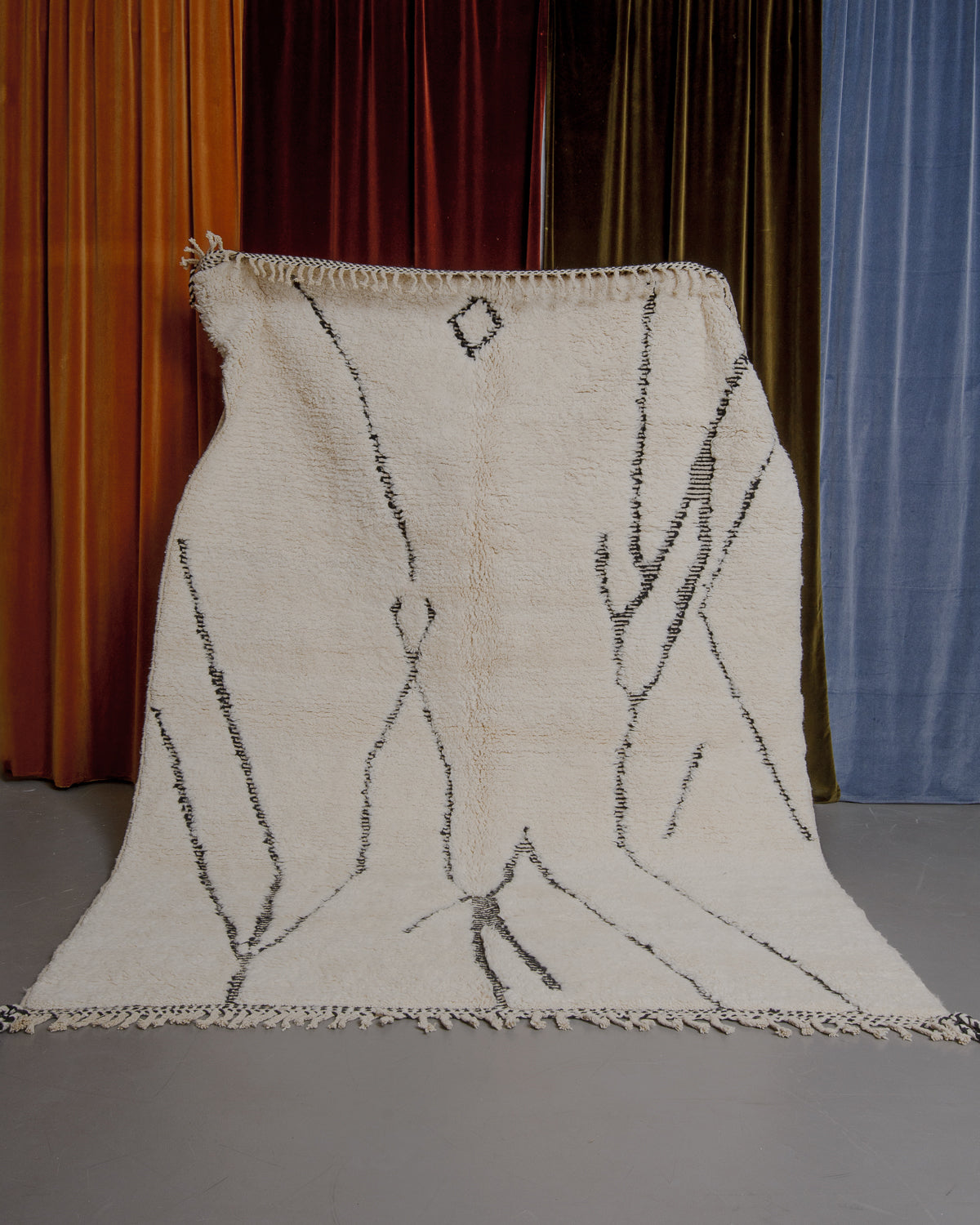 Cream and black Berberlin Beniourain rug held up in front of velvet curtains.