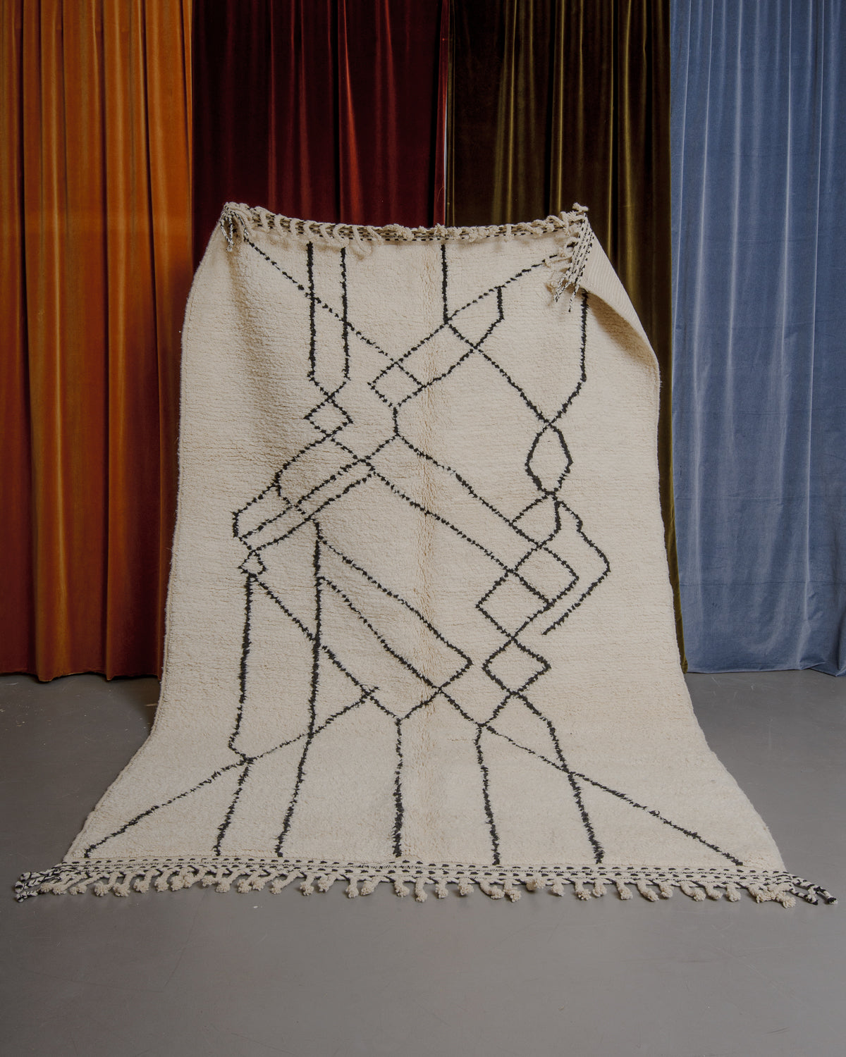 Cream and black Berberlin Beniourain rug held up in front of velvet curtains.
