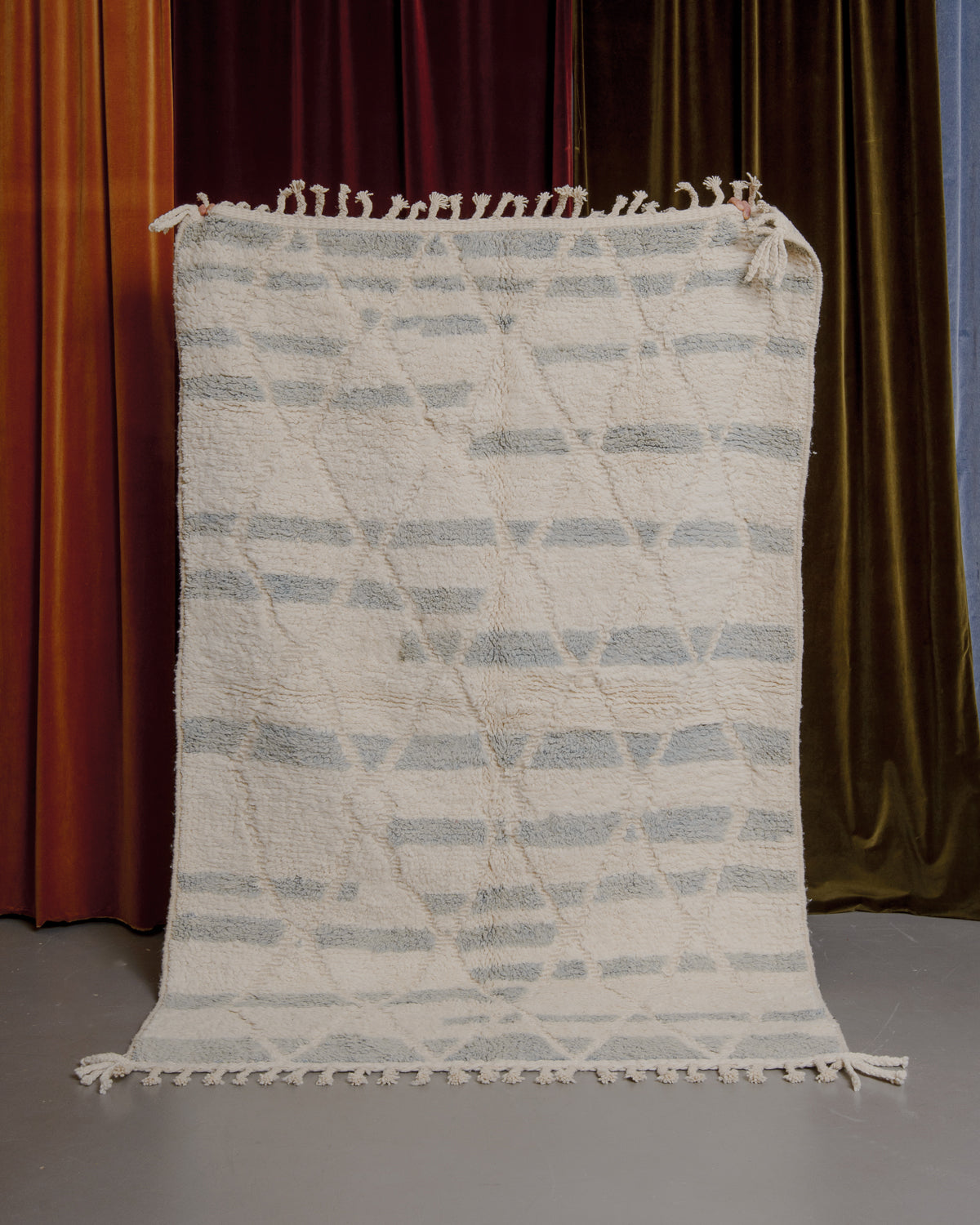 Cream and light blue-grey Berberlin Beniourain rug held up in front of velvet curtains.