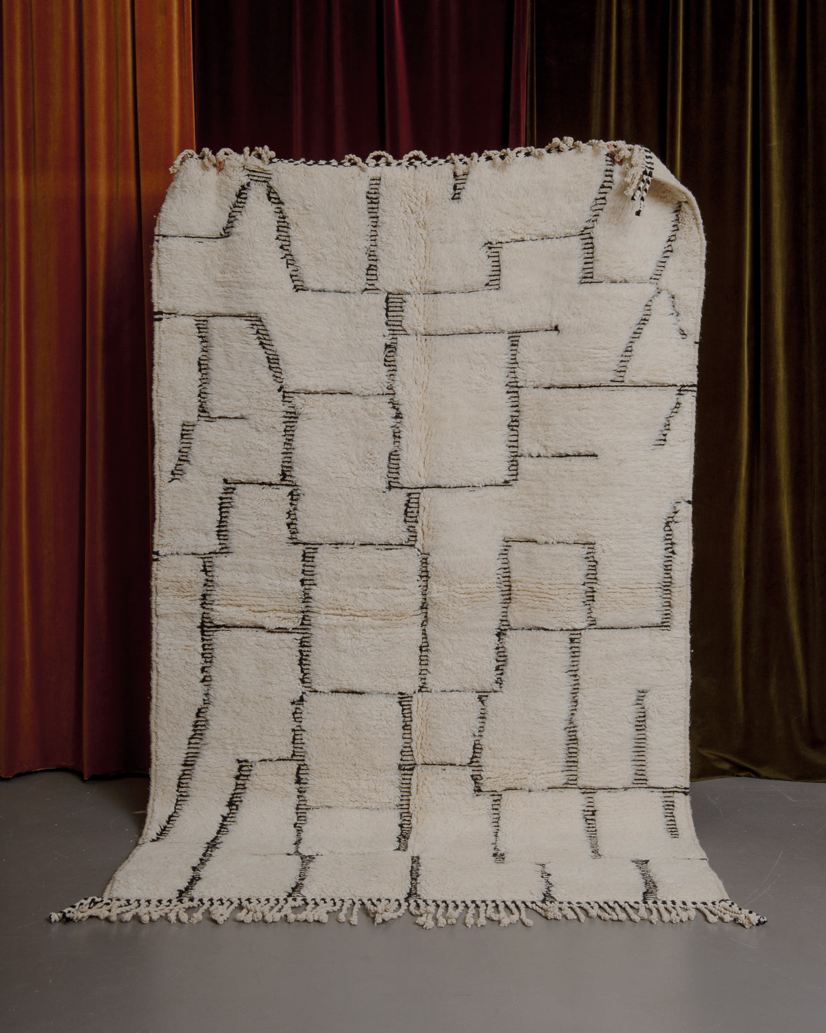 Cream and black Berberlin Beniourain rug held up in front of velvet curtains.