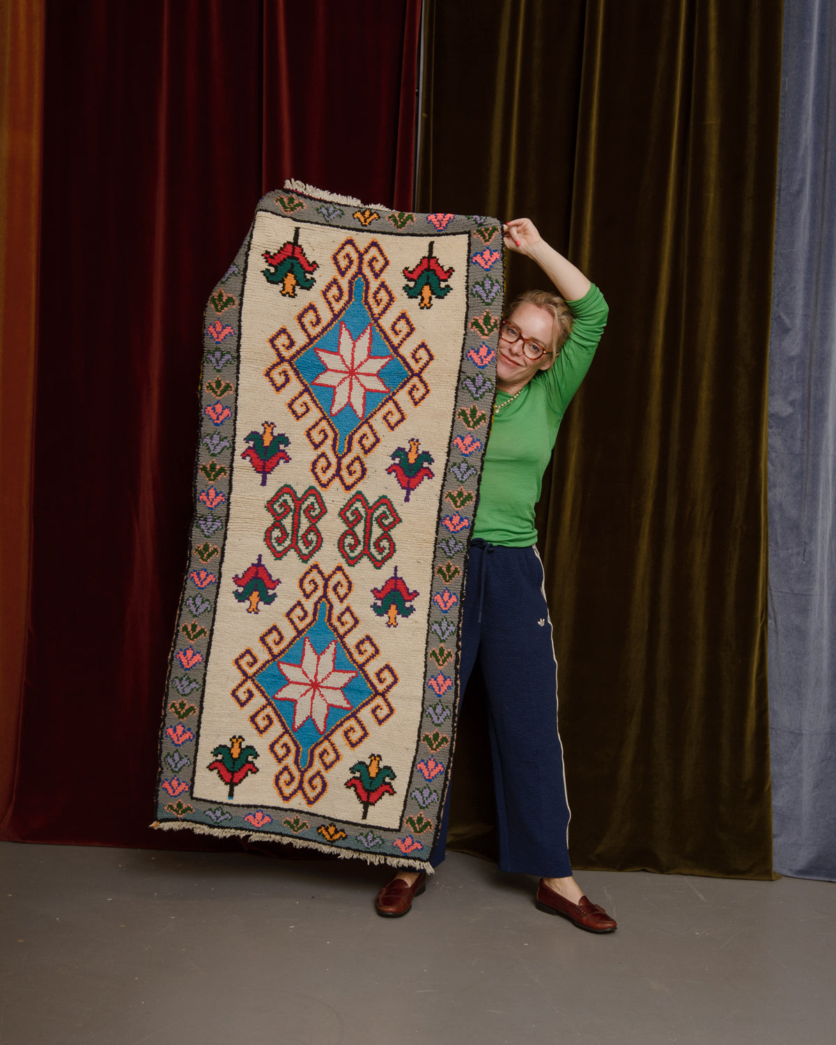 Vintage Berberlin wool runner rug being held up in front of velvet curtains by a woman in a green top and glasses.