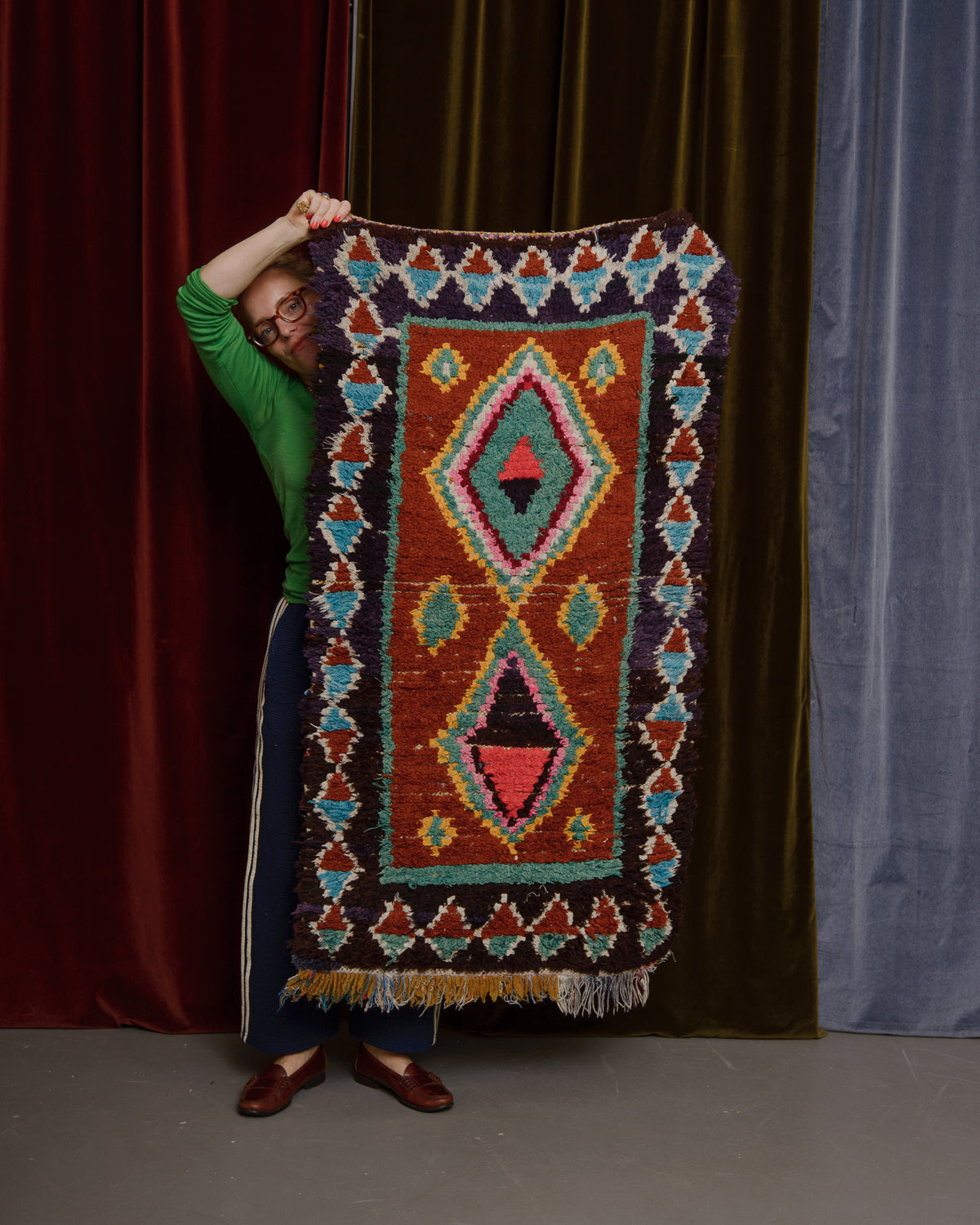 Vintage Berberlin wool runner rug being held up in front of velvet curtains by a woman in brown loafers and glasses.