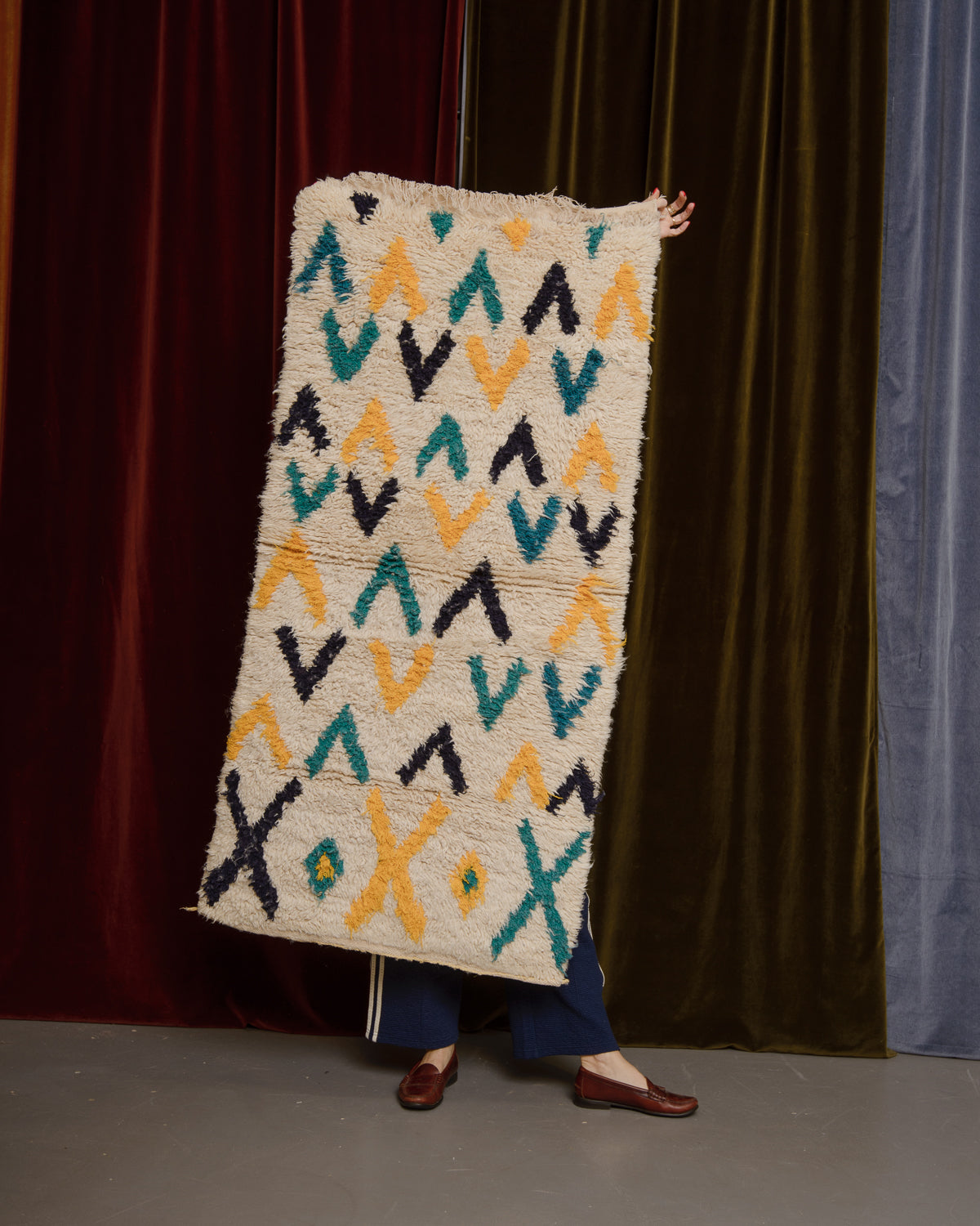Vintage Berberlin wool runner rug being held up in front of velvet curtains by a woman in brown loafers.