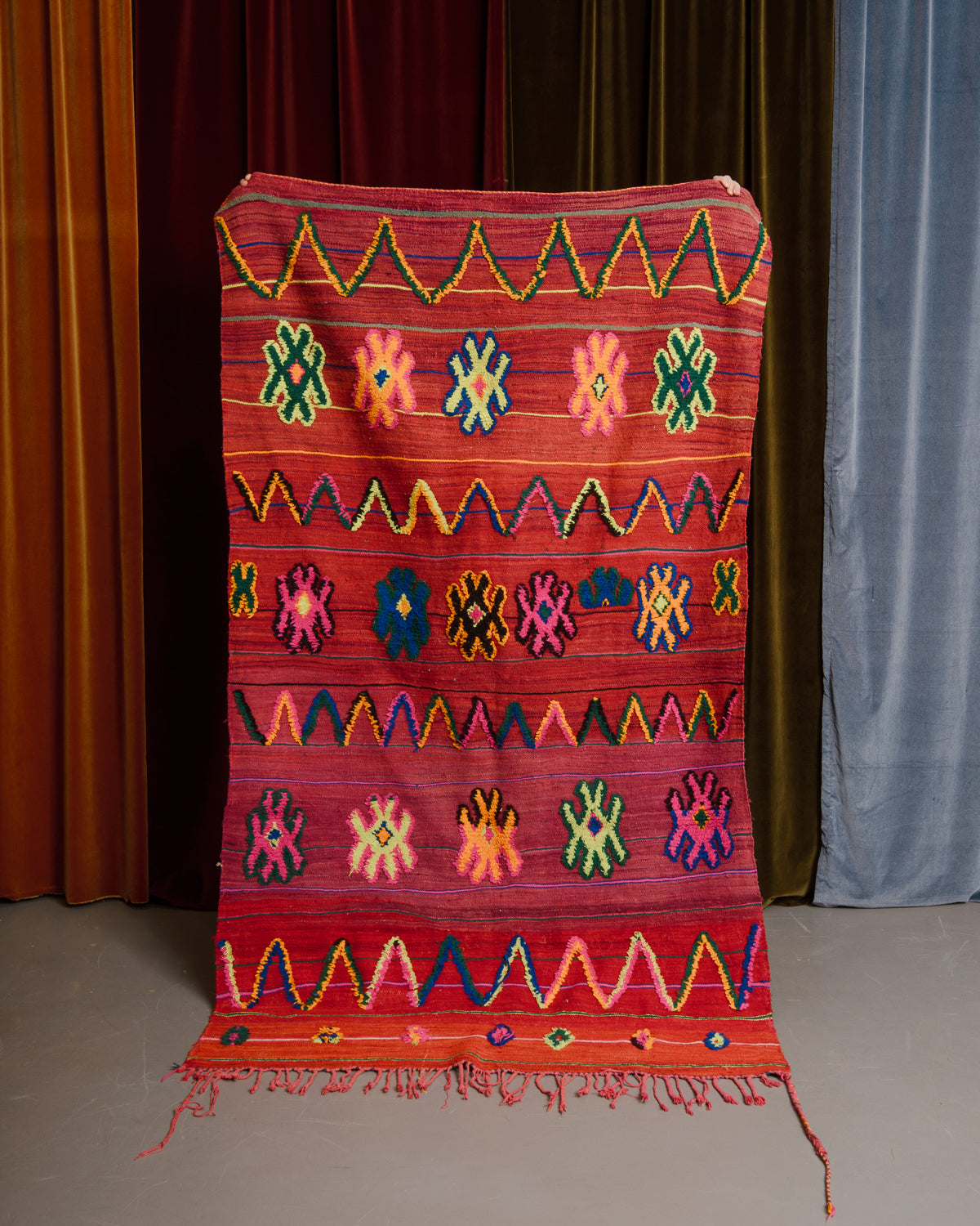 Vintage flat woven Kelim with fluffy embroidered pattern, held in front of velvet curtains. 