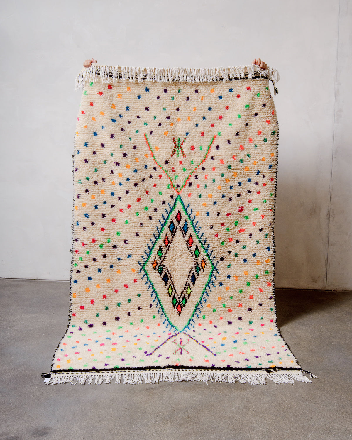Berberlin Azilal rug made of cream sheep’s wool and colourful cotton with hands holding it up in front of a white studio wall.