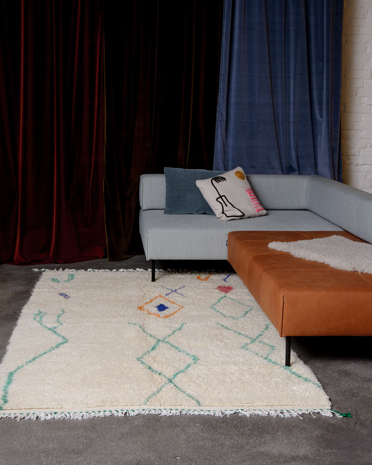 Cream wool Azilal rug with symbols and patterns in turquoise, blue, orange and pink, laying on concrete floor under modular couch with velvet curtains behind.