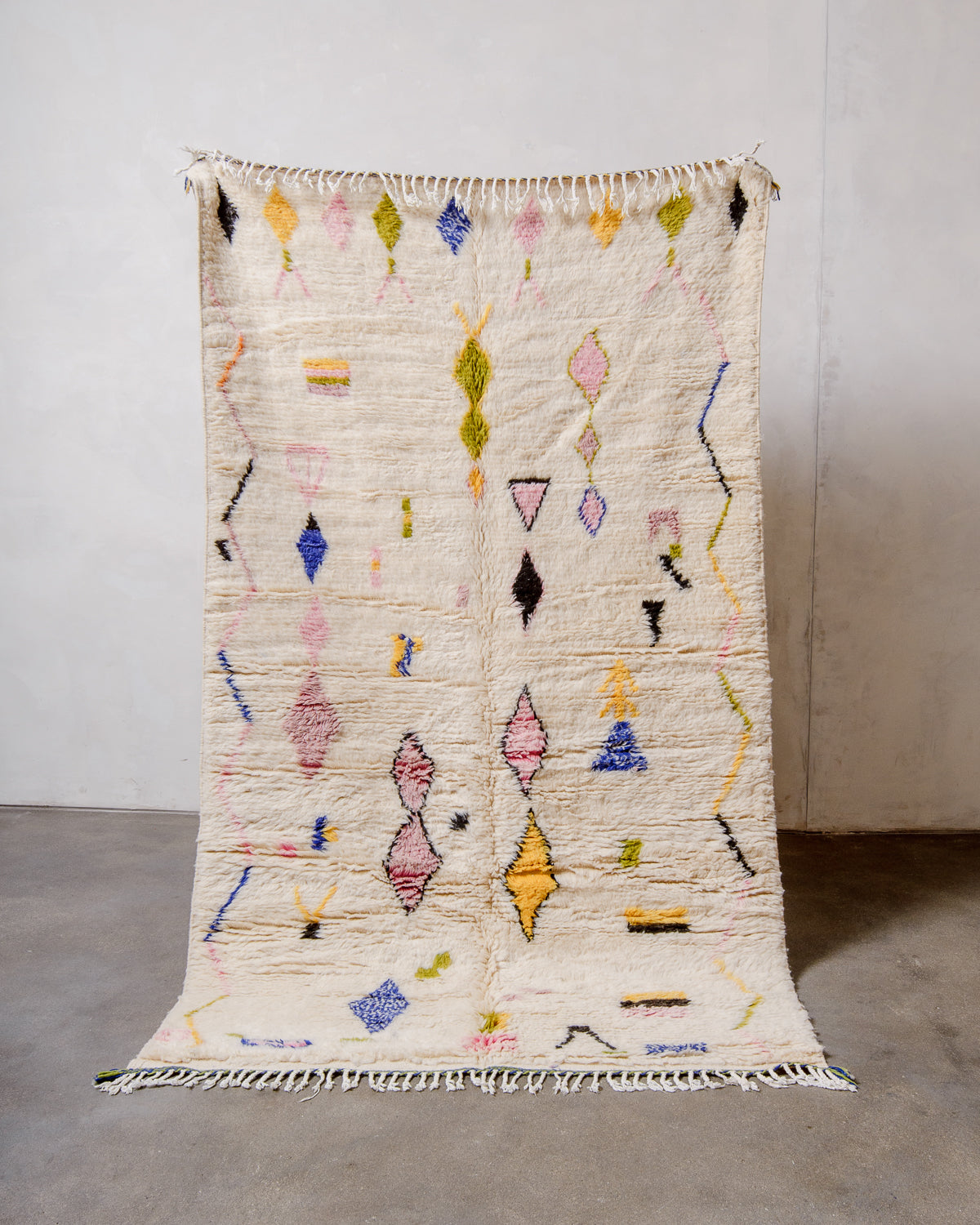 Berberlin Azilal rug in fluffy cream wool with colourful symbols and shapes, being held up in front of a white wall.