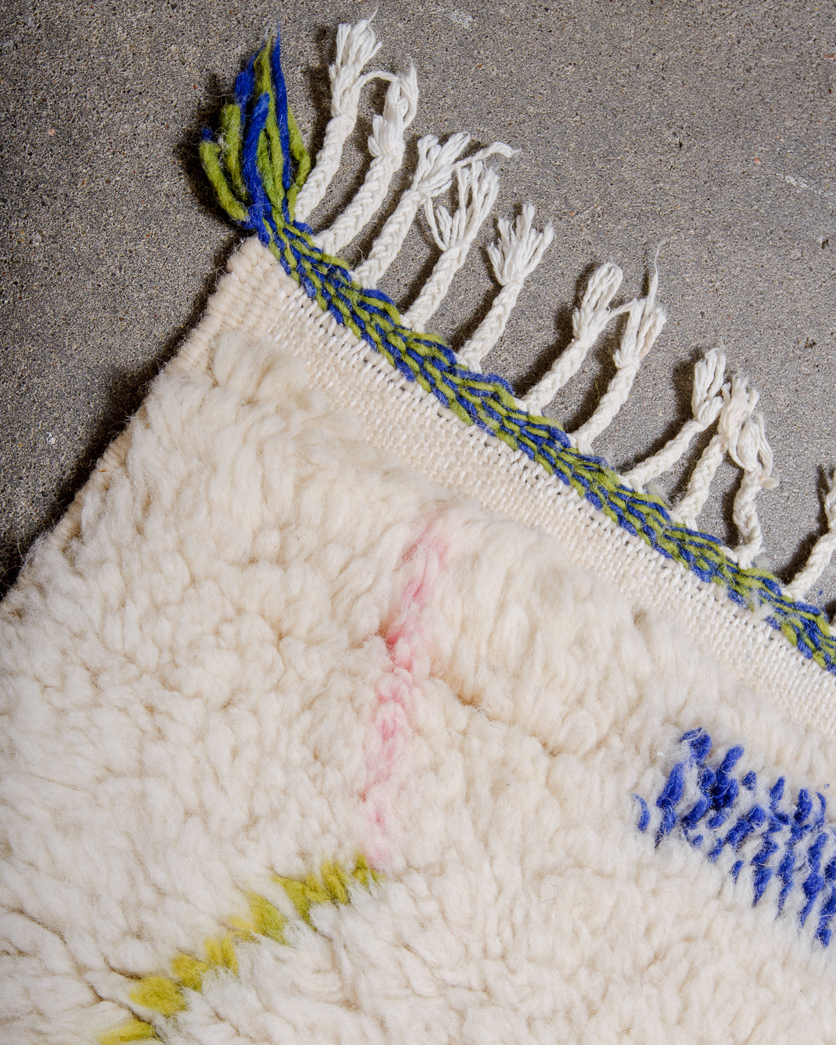 Corner of cream Azilal rug with green and blue woven edge and white cotton tassels. 