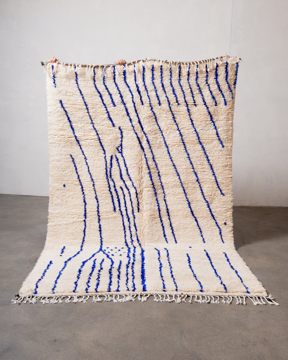 Berberlin Azilal rug made of fluffy cream wool and design of abstract blue lines, held up against a white wall and cement floor.
