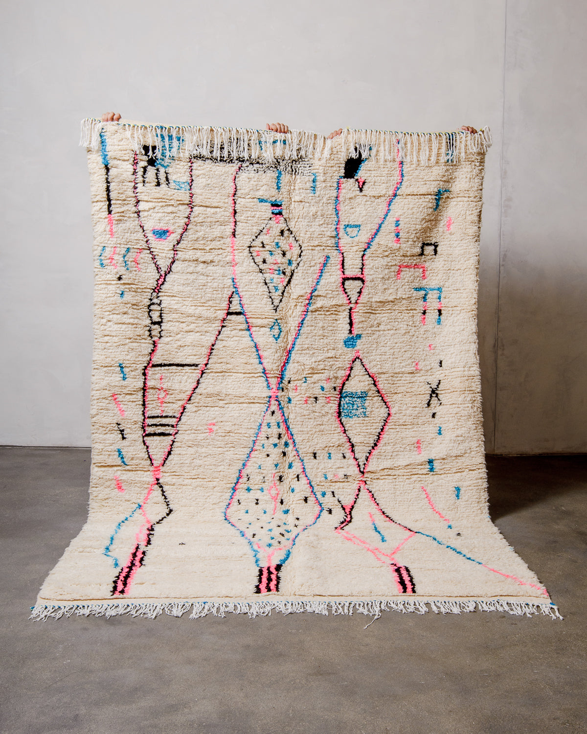 Berberlin Azilal rug made of fluffy cream wool and colourful design, held up  against a white wall and cement floor.