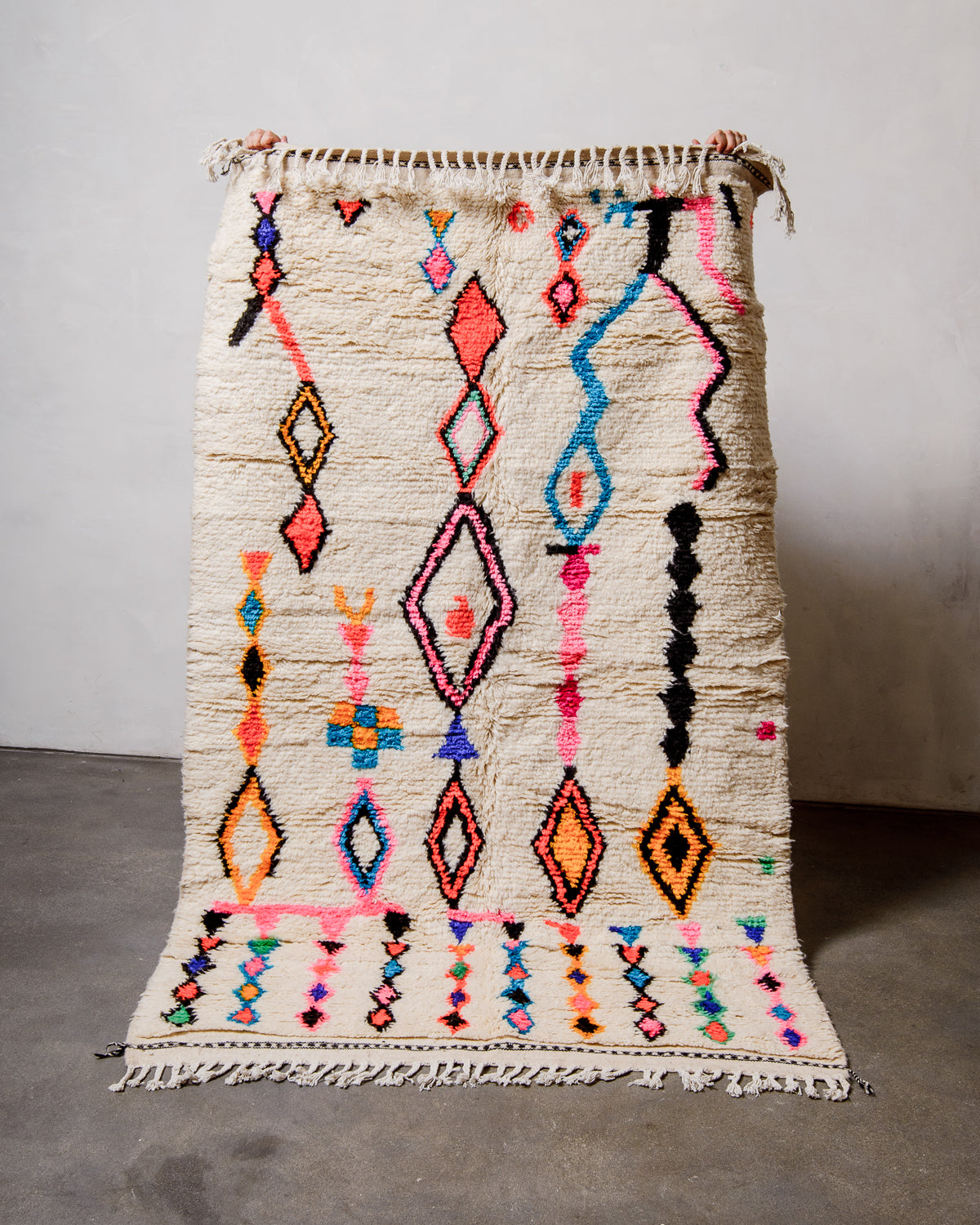 Berberlin Azilal rug made of fluffy cream wool and colourful design, held up  against a white wall and cement floor.