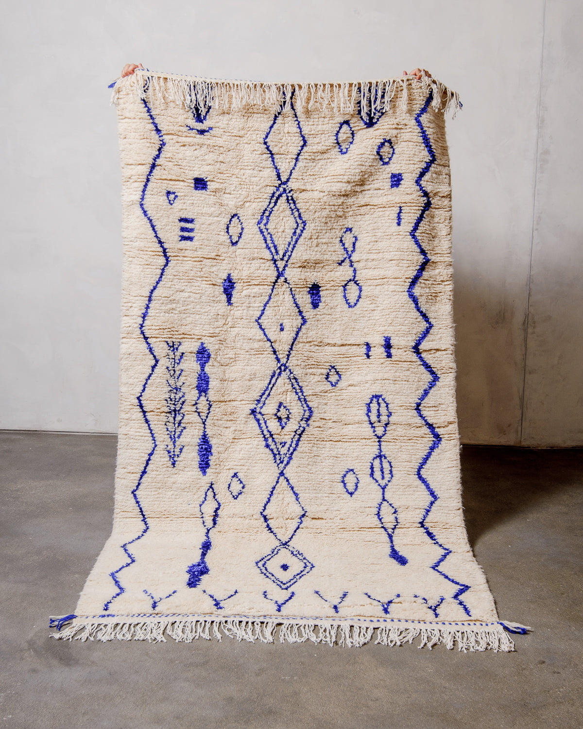 Berberlin Azilal rug made of fluffy cream wool and bright blue cotton, held up  against a white wall and cement floor.