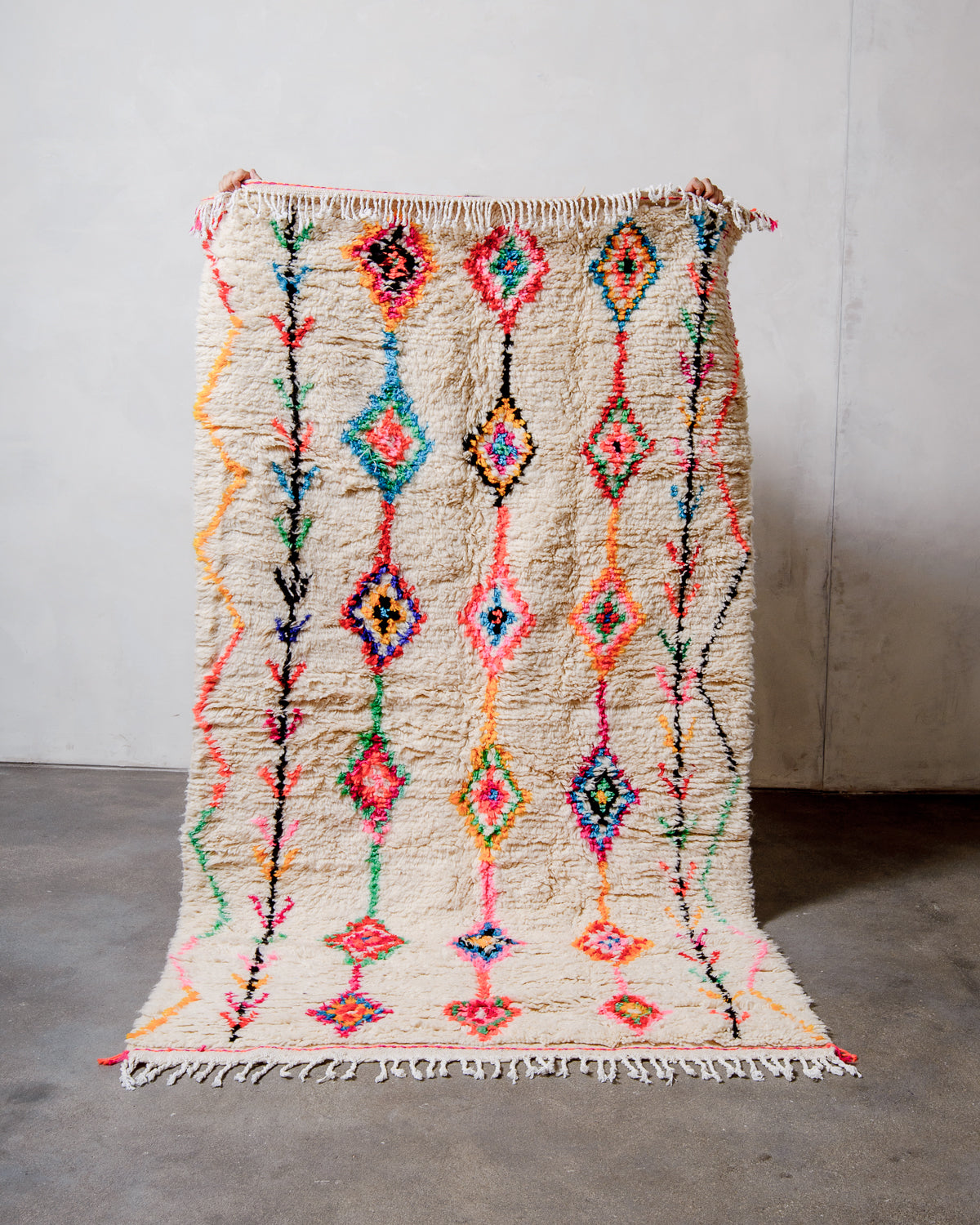 Berberlin Azilal rug made of fluffy cream wool and a brightly coloured pattern with cotton tassels, held up  against a white wall and cement floor.