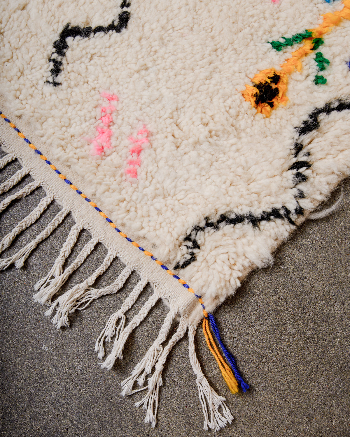 Corner of cream Azilal rug with white cotton tassels across the end and blue and yellow tassels at the corner.  