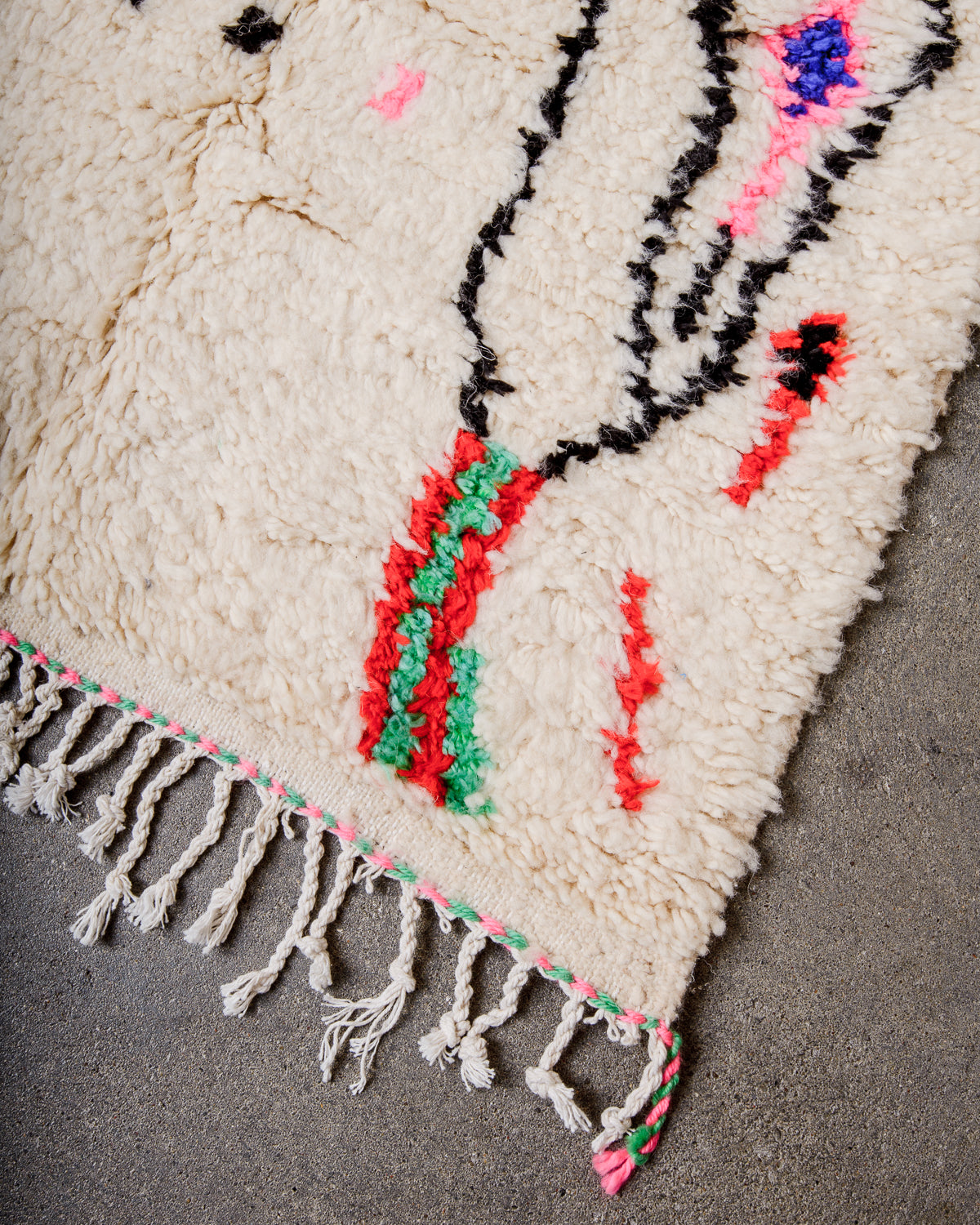 Corner of cream Azilal rug with white cotton tassels across the end and pink and green tassels at the corner.  