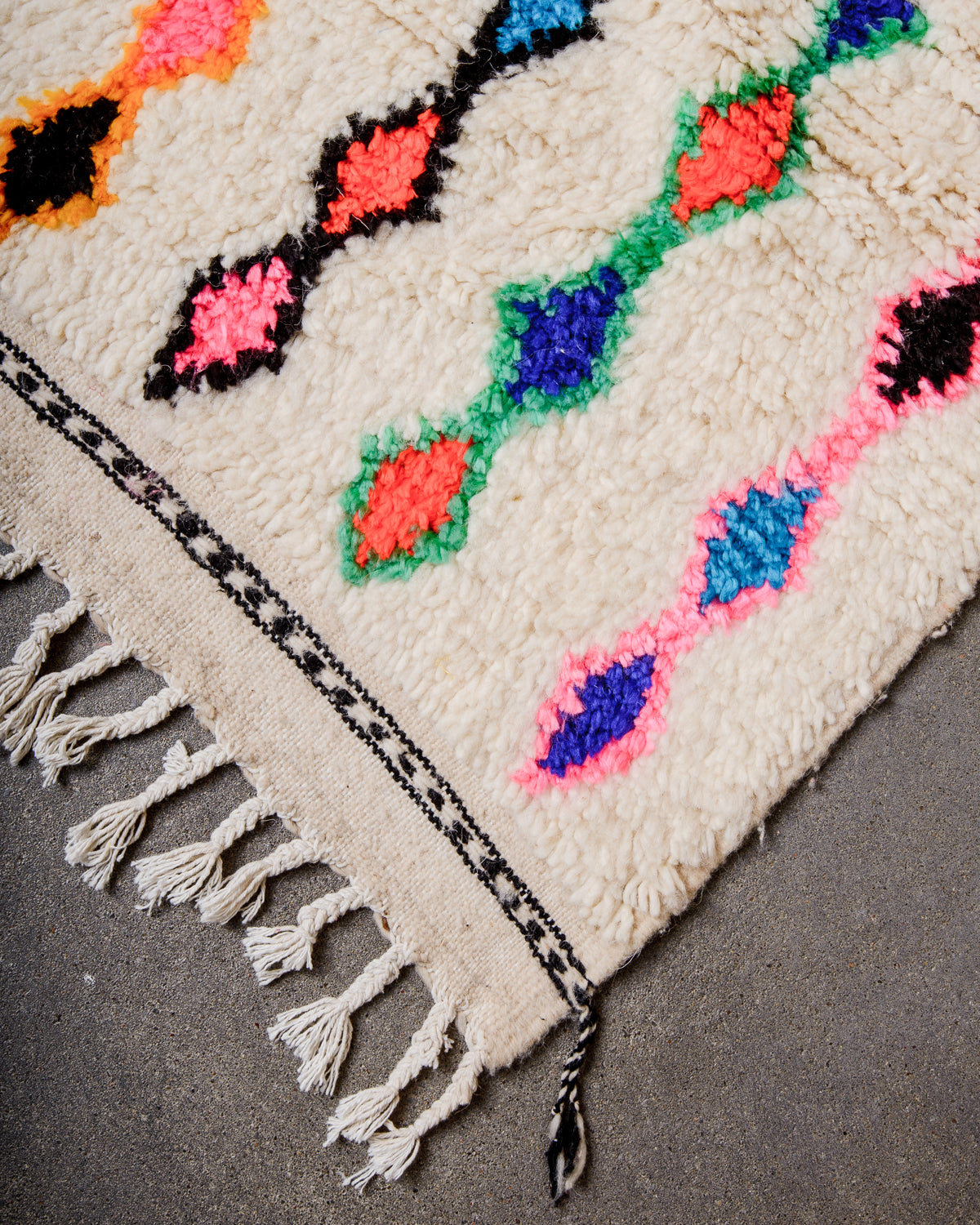 Corner of cream Azilal rug with white cotton tassels across the top and a black embroidered design across the tightly woven end of the rug.