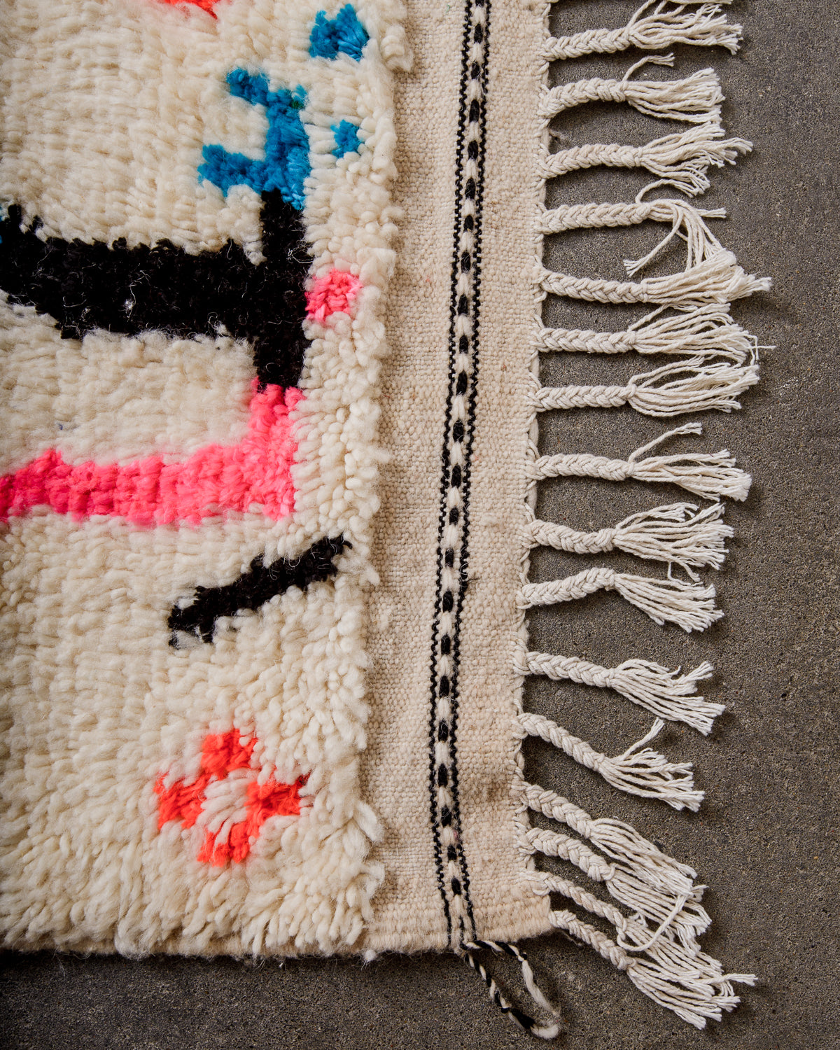 Corner of cream Azilal rug with white cotton tassels across the top and a black embroidered design across the tightly woven end of the rug.