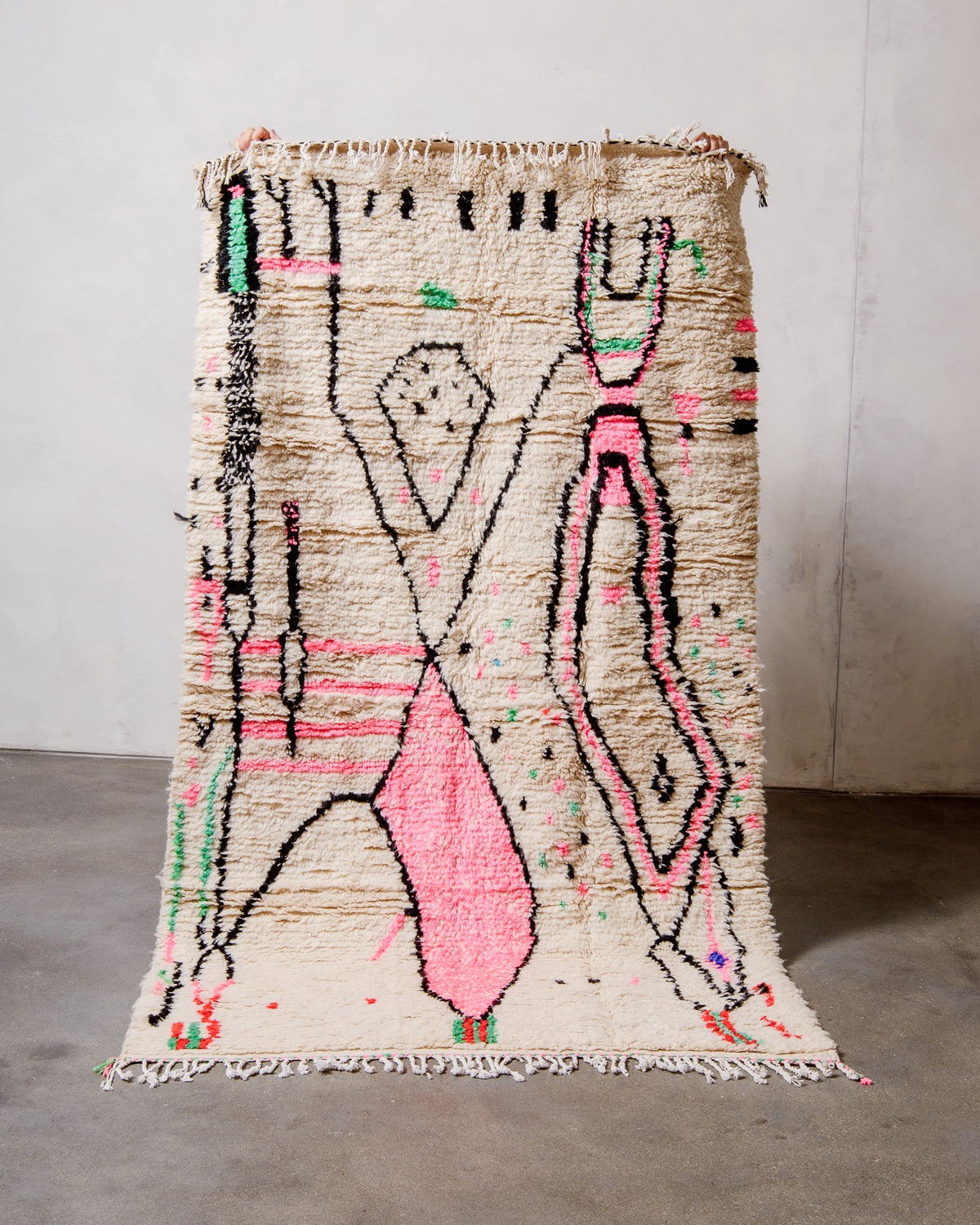 Berberlin Azilal rug made of fluffy cream wool with cotton tassels, held up  against a white wall and cement floor.