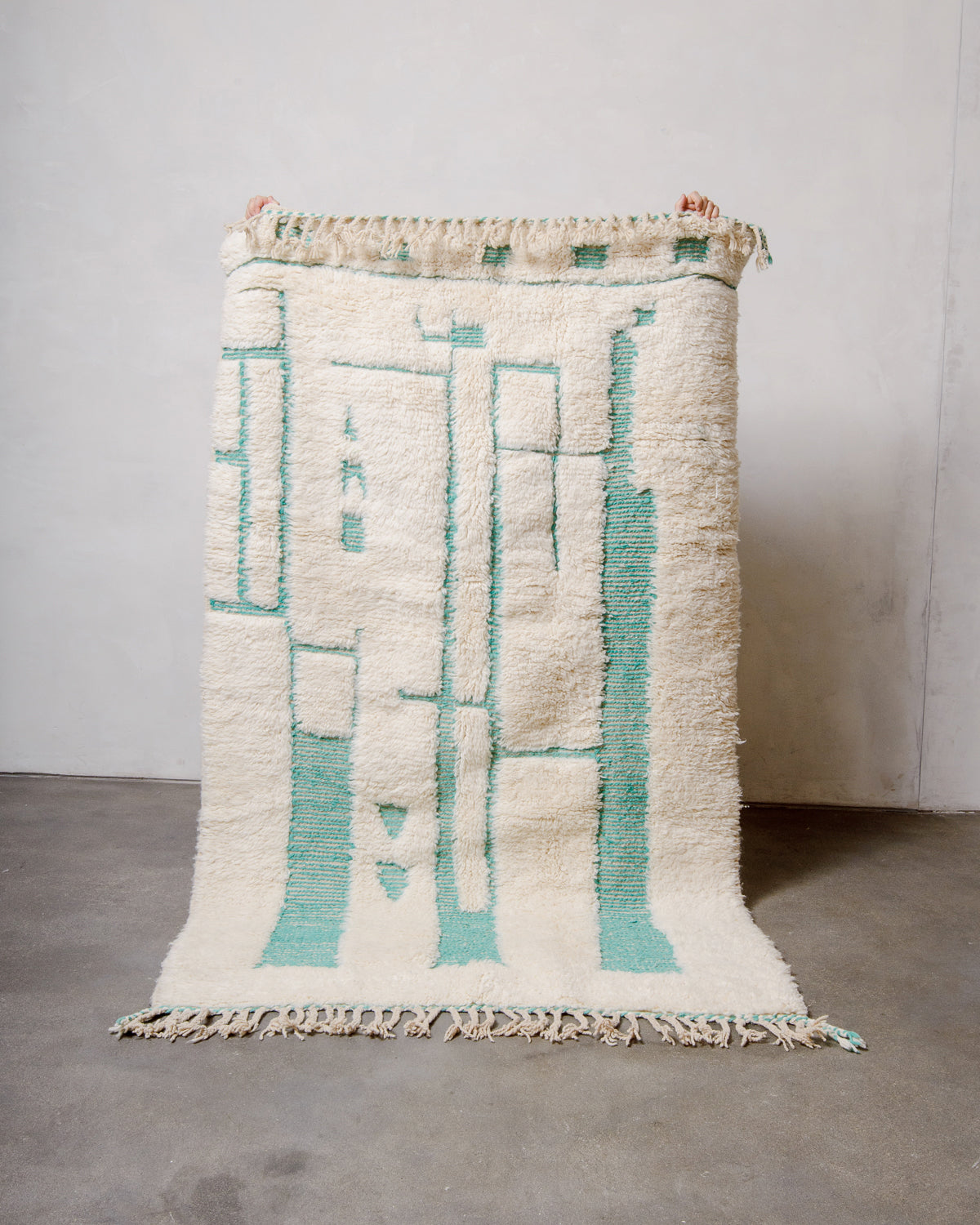 Berberlin Beniourain rug in cream and turquoise wool being held up against a white wall and a concrete floor. 
