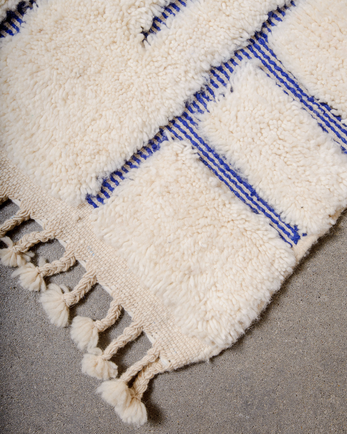 Corner of cream Berber rug with natural wool tassels laying on concrete floor. 