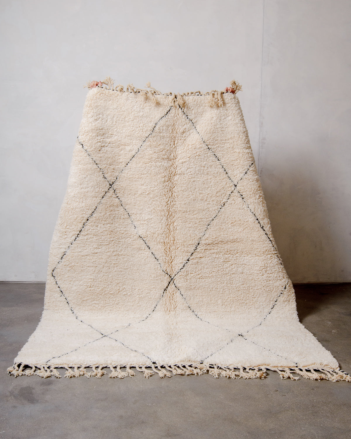 Cream wool Beniourain rug with minimal black line design being held up in front of a white wall and concrete floor. 