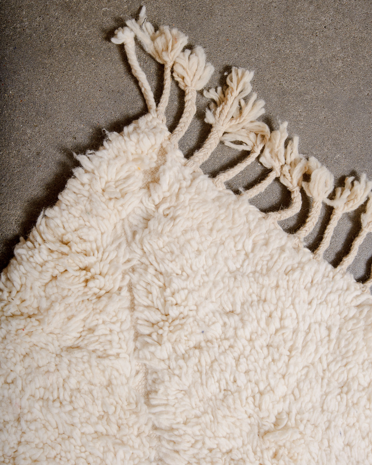 Corner of fluffy cream wool Beniourain rug with natural wool tassels laying on concrete floor. 