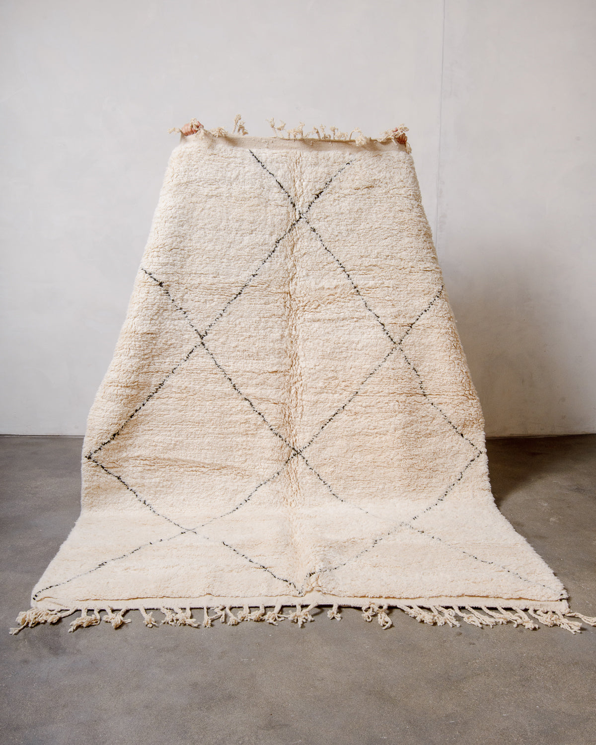 Cream wool Beniourain rug with minimal black line design being held up in front of a white wall and concrete floor. 
