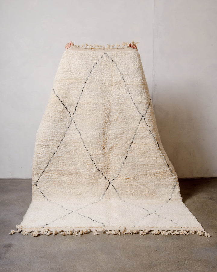 Cream Berber rug with black diamond design being held up by two hands in front of white wall. 