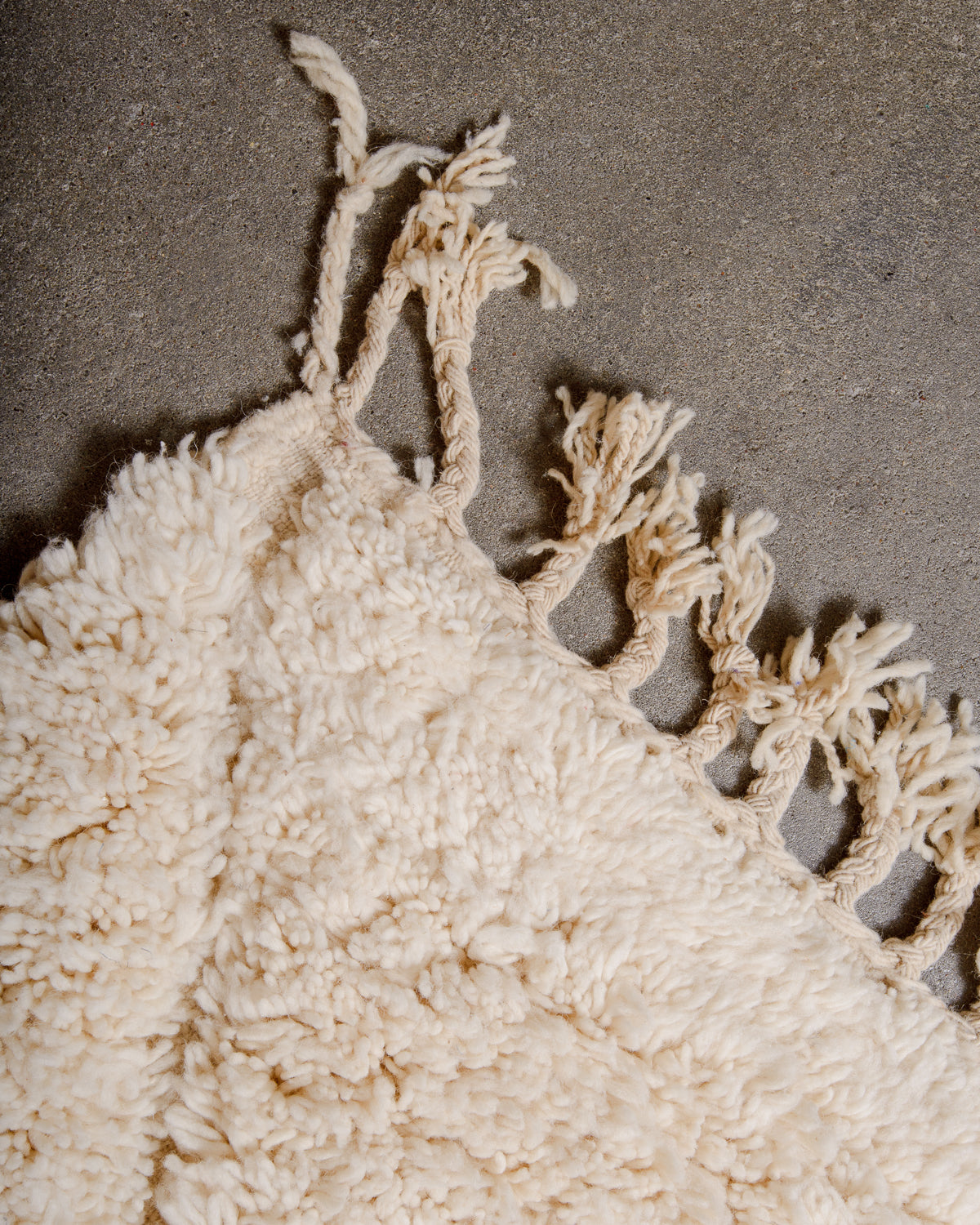 Corner of fluffy cream wool Beniourain rug with natural wool tassels laying on concrete floor. 