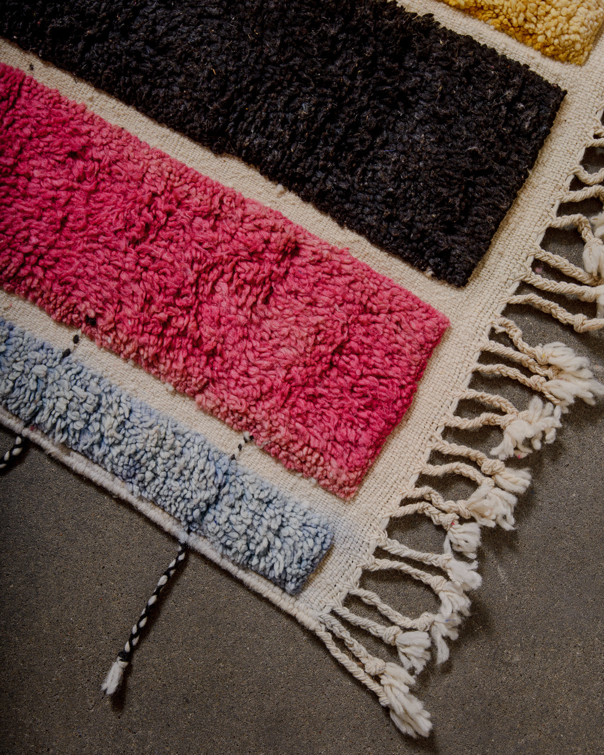 Corner of Beniourain rug with knotted wool in light blue, pink, black and yellow and natural wool tassels on the end. There are black and white tassels also on the sides. 