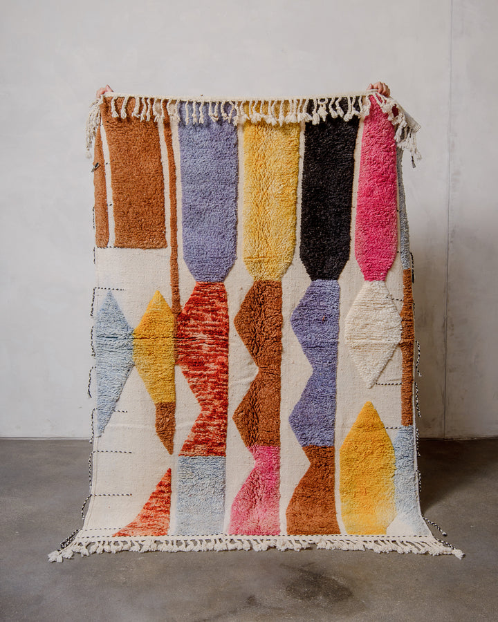 Berberlin Beniourain rug in colourful wool and cream, being held up in front of a white wall. 