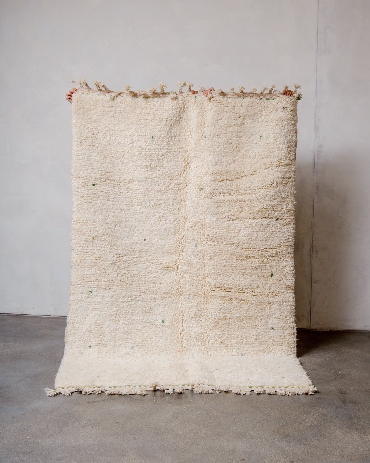 Berberlin Beniourain rug in cream wool with coloured dots being held up in front of a white wall. 