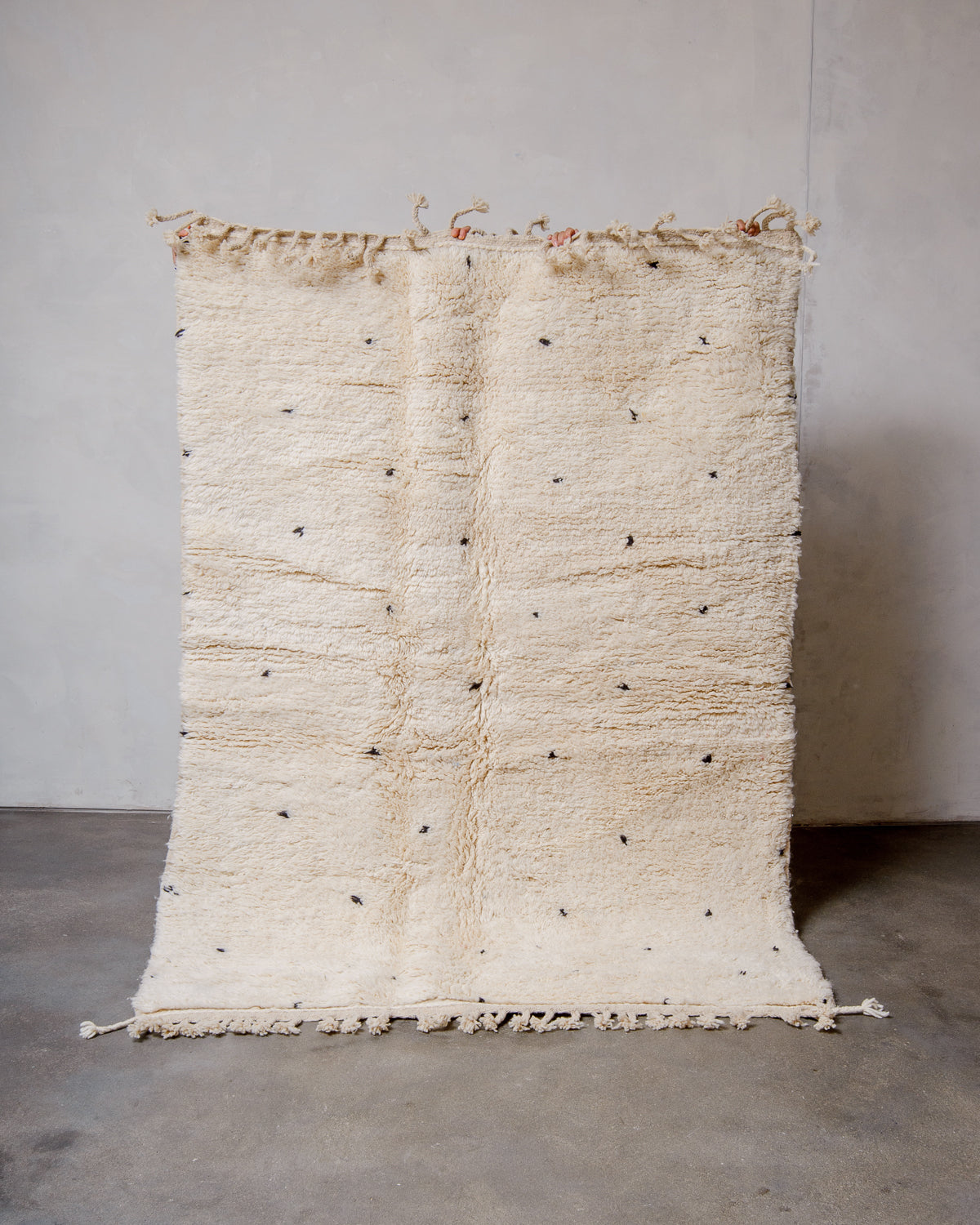 Berberlin Beniourain rug in cream wool with black dots being held up in front of a white wall. 