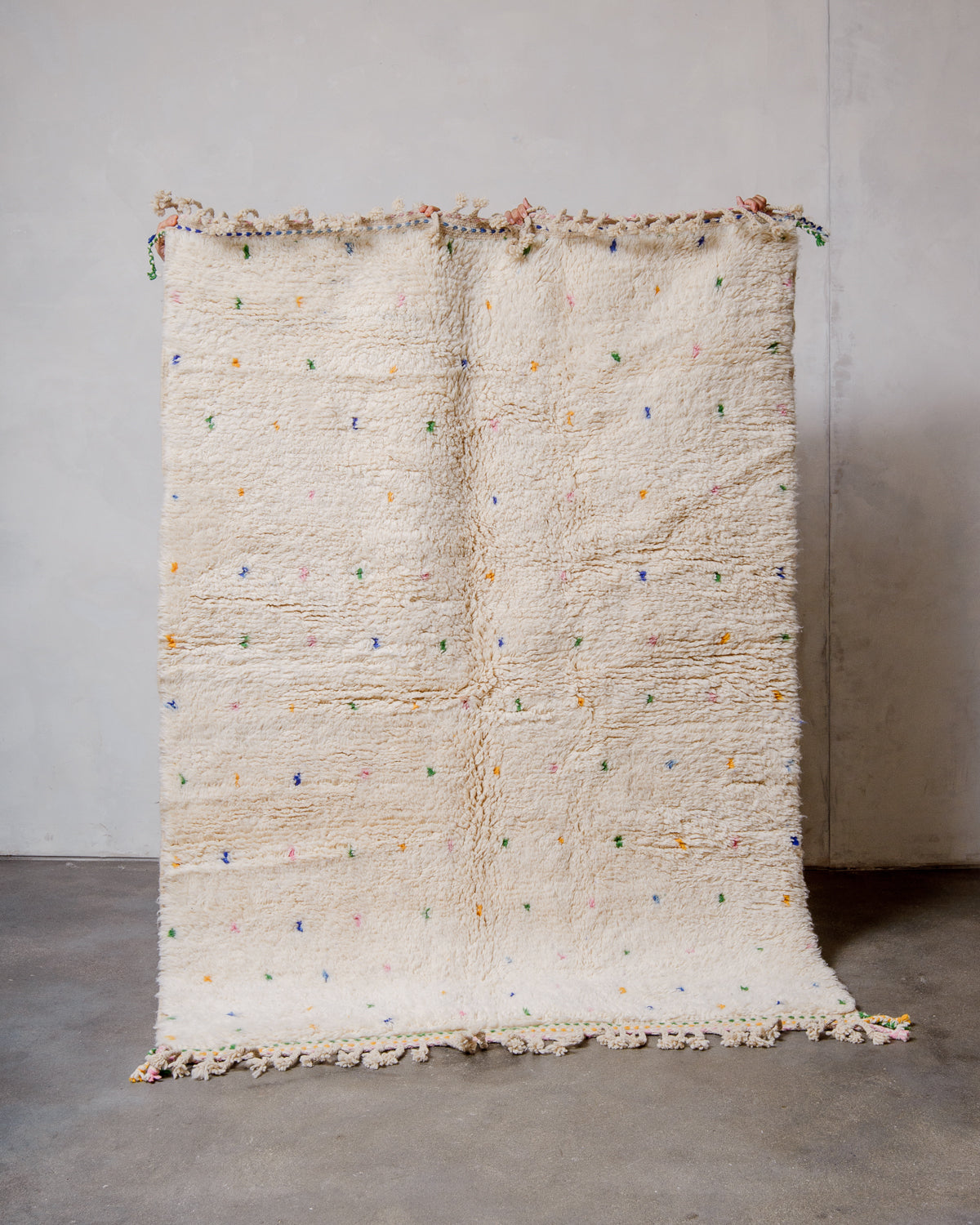 Berberlin Beniourain rug in cream wool with coloured dots being held up in front of a white wall. 