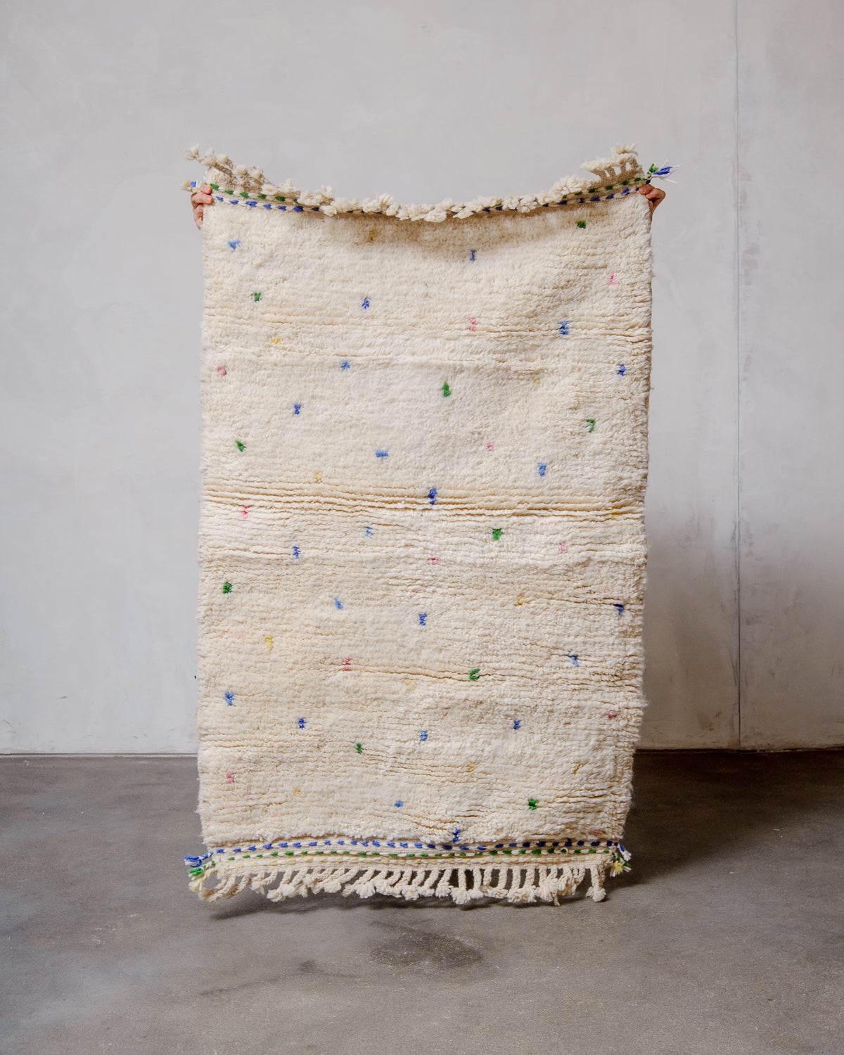 Cream Berber rug with coloured dots being held up in front of a white wall. 
